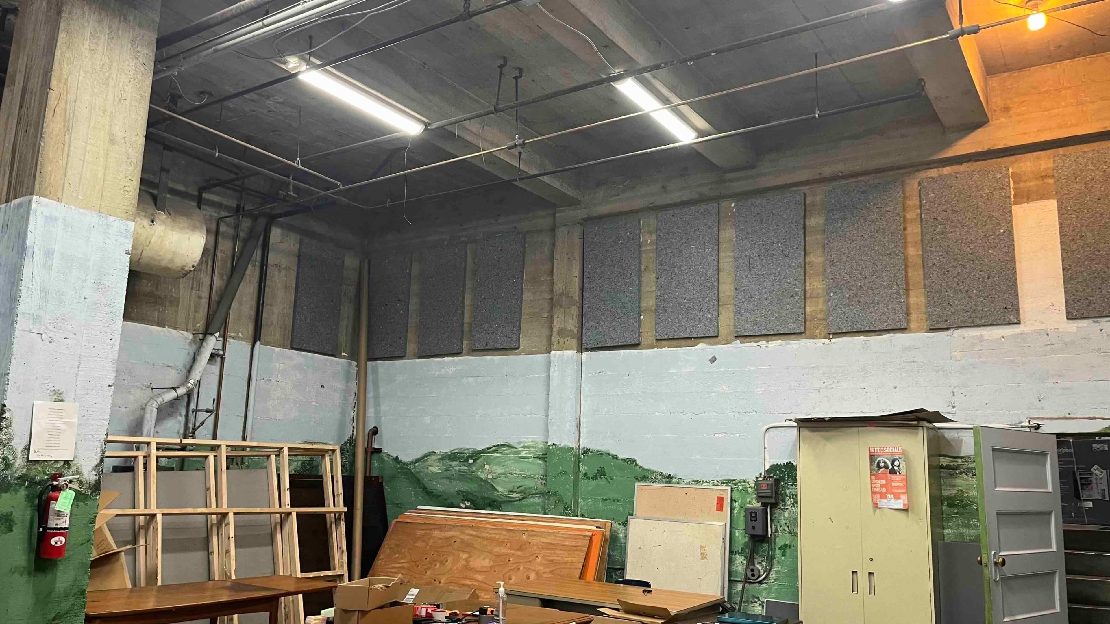 The image depicts a dimly lit industrial room with exposed concrete walls, wooden furniture stacked against the wall, and soundproofing panels installed above.