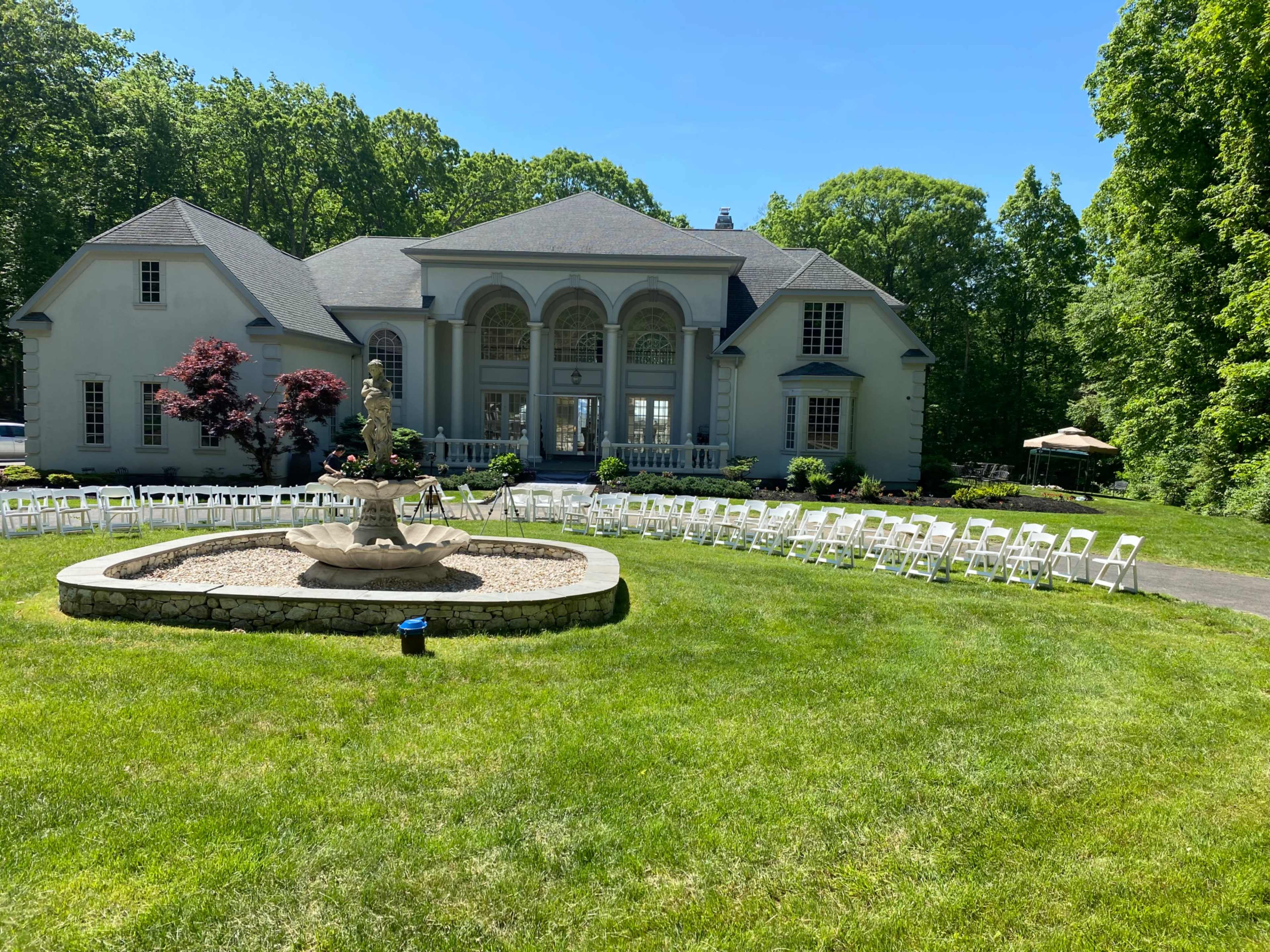 A spacious outdoor venue features a large house with arched windows, a central fountain surrounded by gravel, and rows of white chairs set up on a grassy lawn.