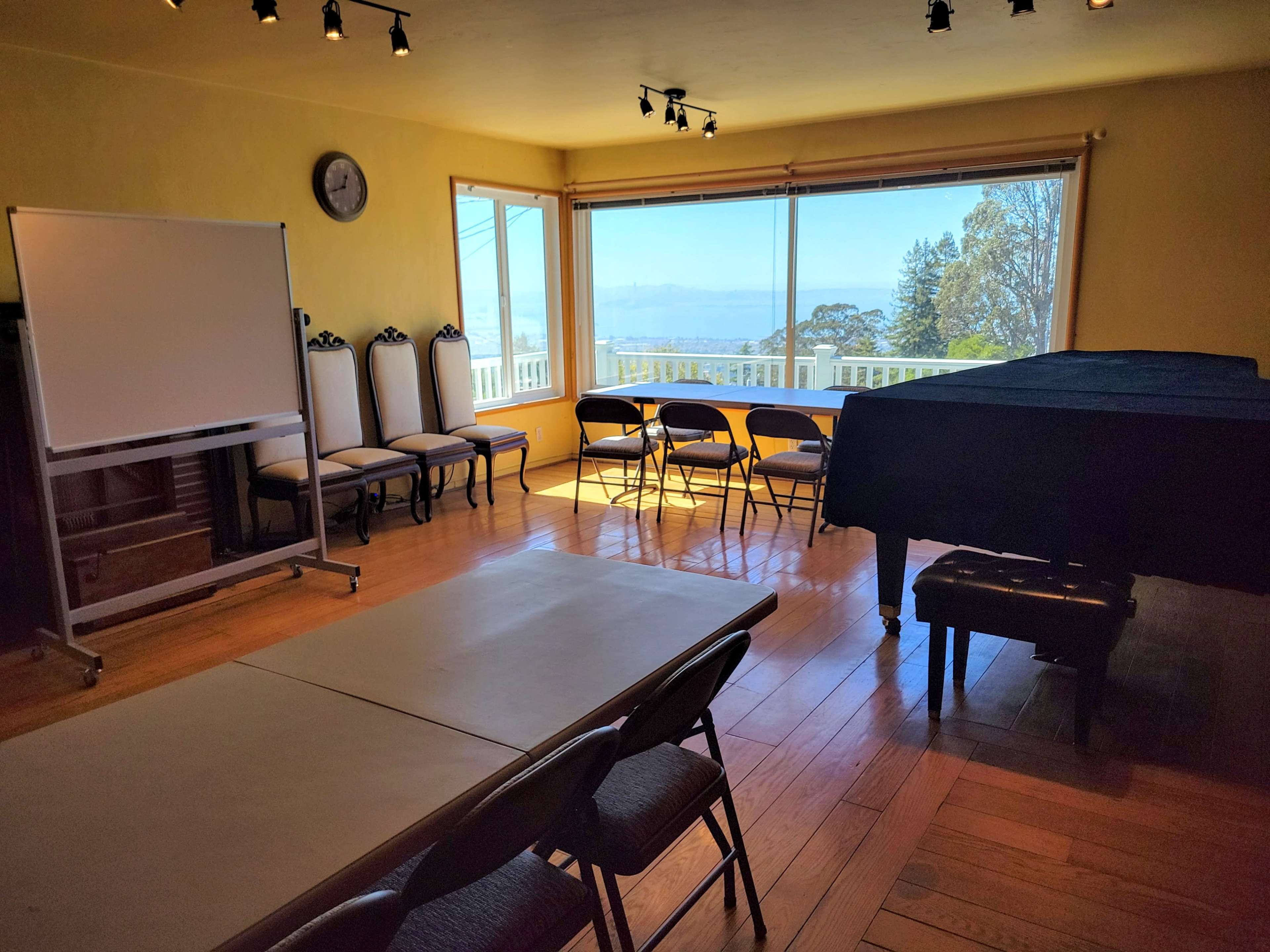 A room with a large window overlooks a view, featuring a piano, a table, chairs, and a whiteboard.