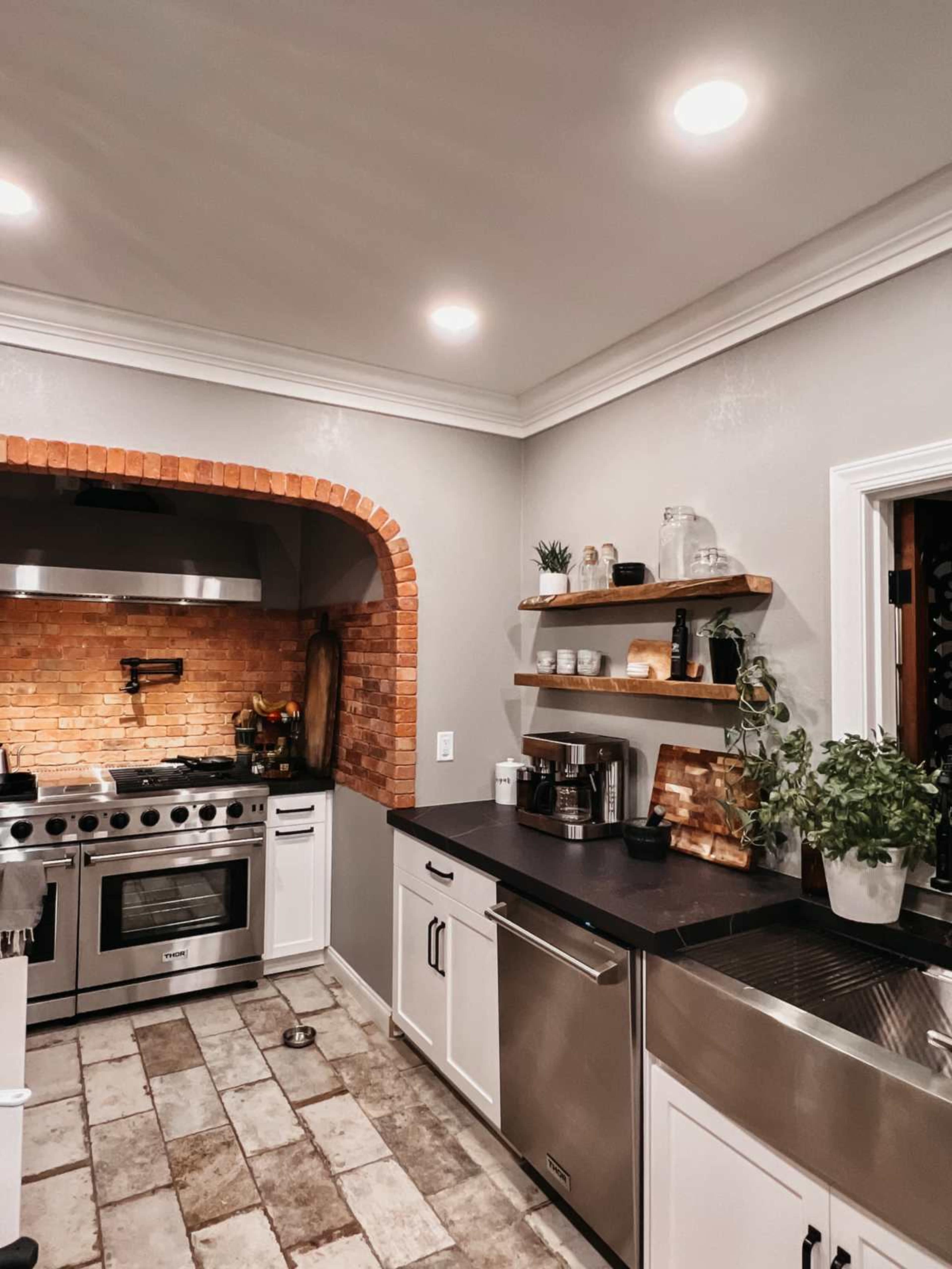 The kitchen features a brick archway over a stainless steel stove, white cabinetry, and open shelves displaying kitchenware.