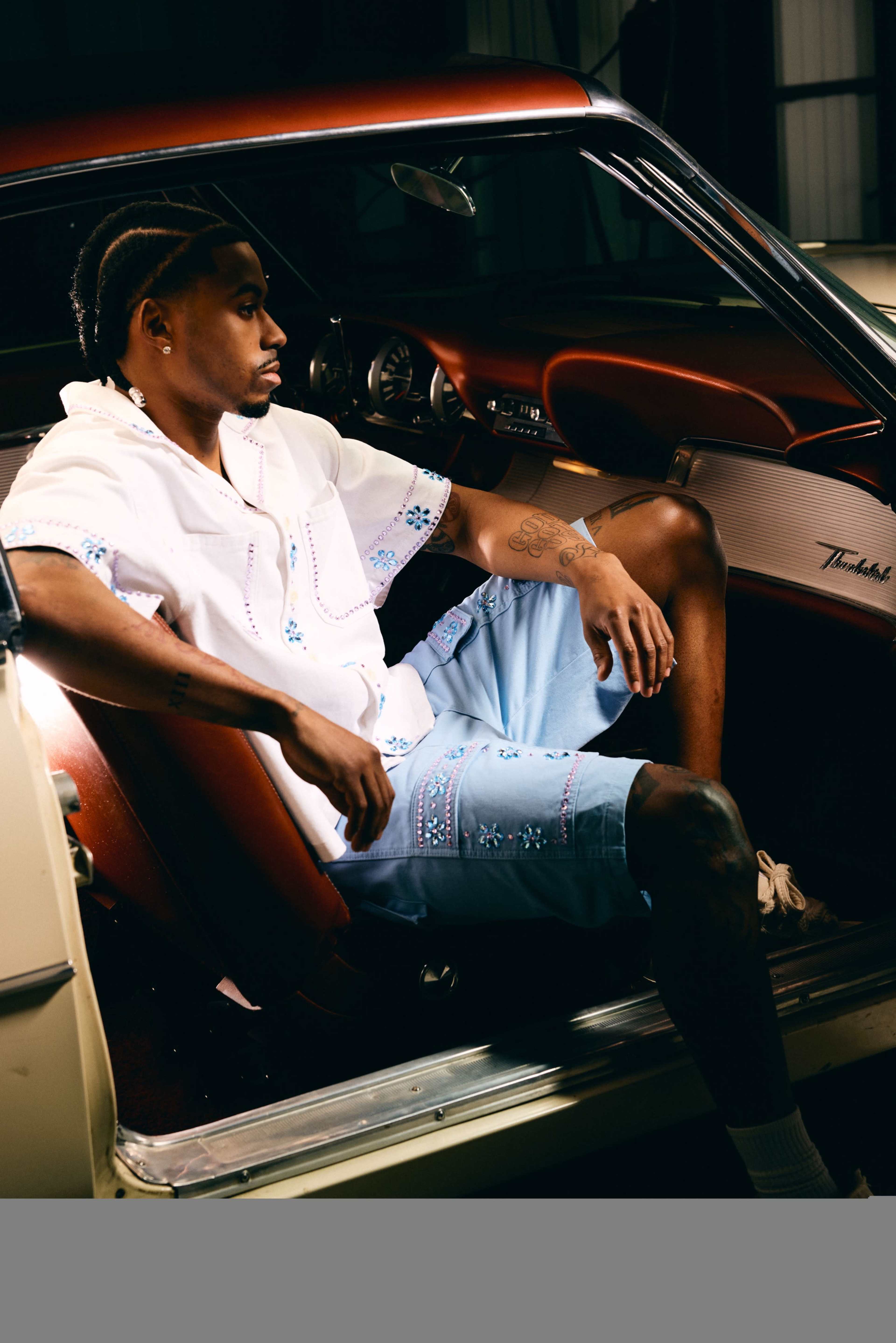 A man in a white embroidered shirt and denim shorts sits in the driver's seat of a vintage car.