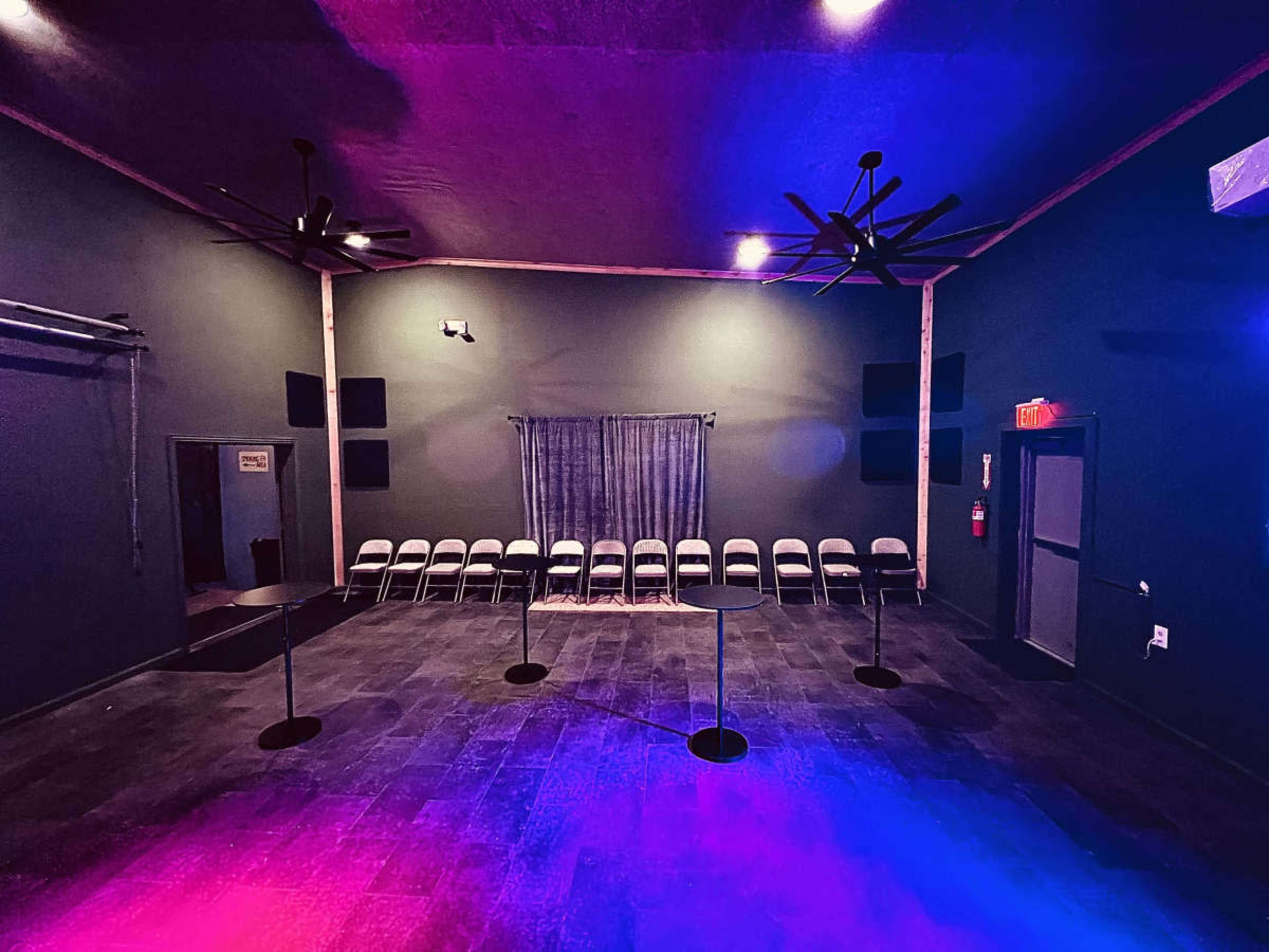 The image shows an empty room setup for an event, featuring a row of chairs facing a curtain, with ceiling fans and colored lighting illuminating the space.