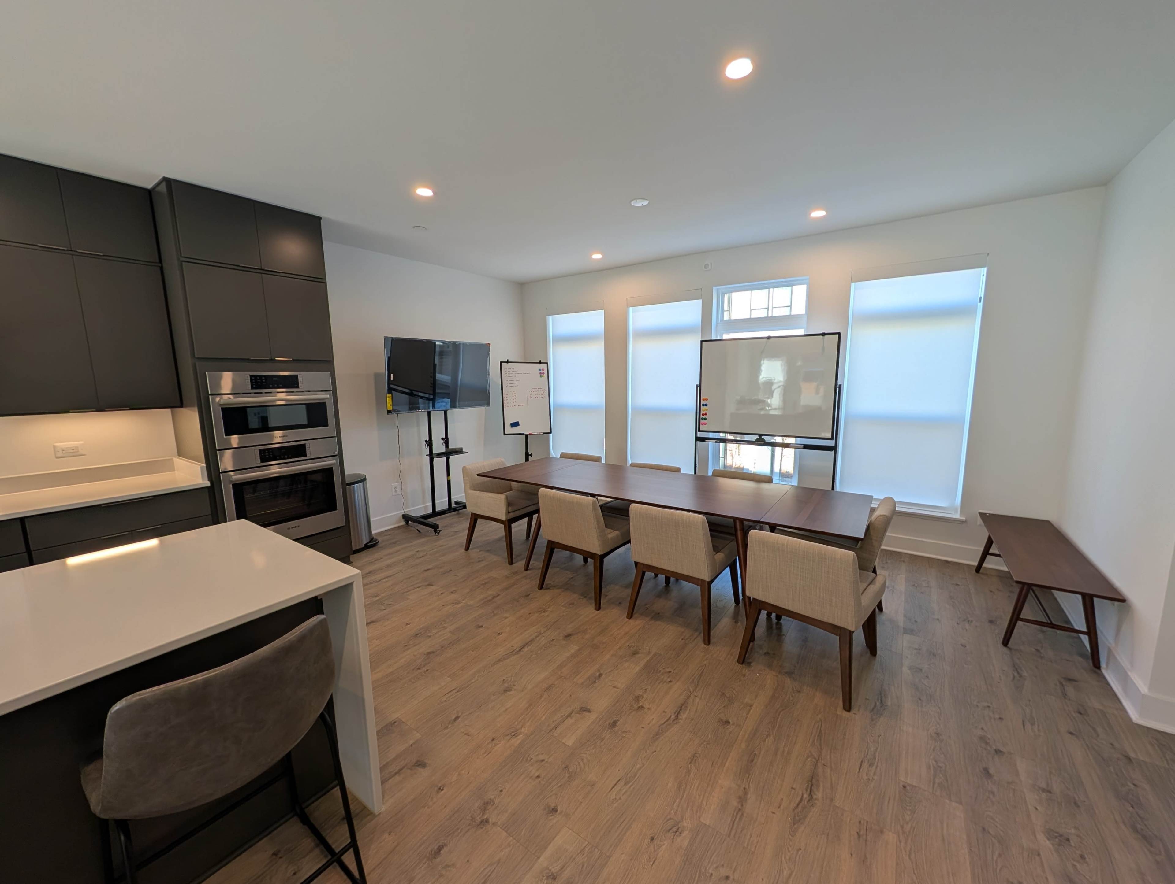 The image shows a modern meeting room with a long wooden table, several chairs, a large whiteboard, and a television against a wall, illuminated by natural light from large windows.