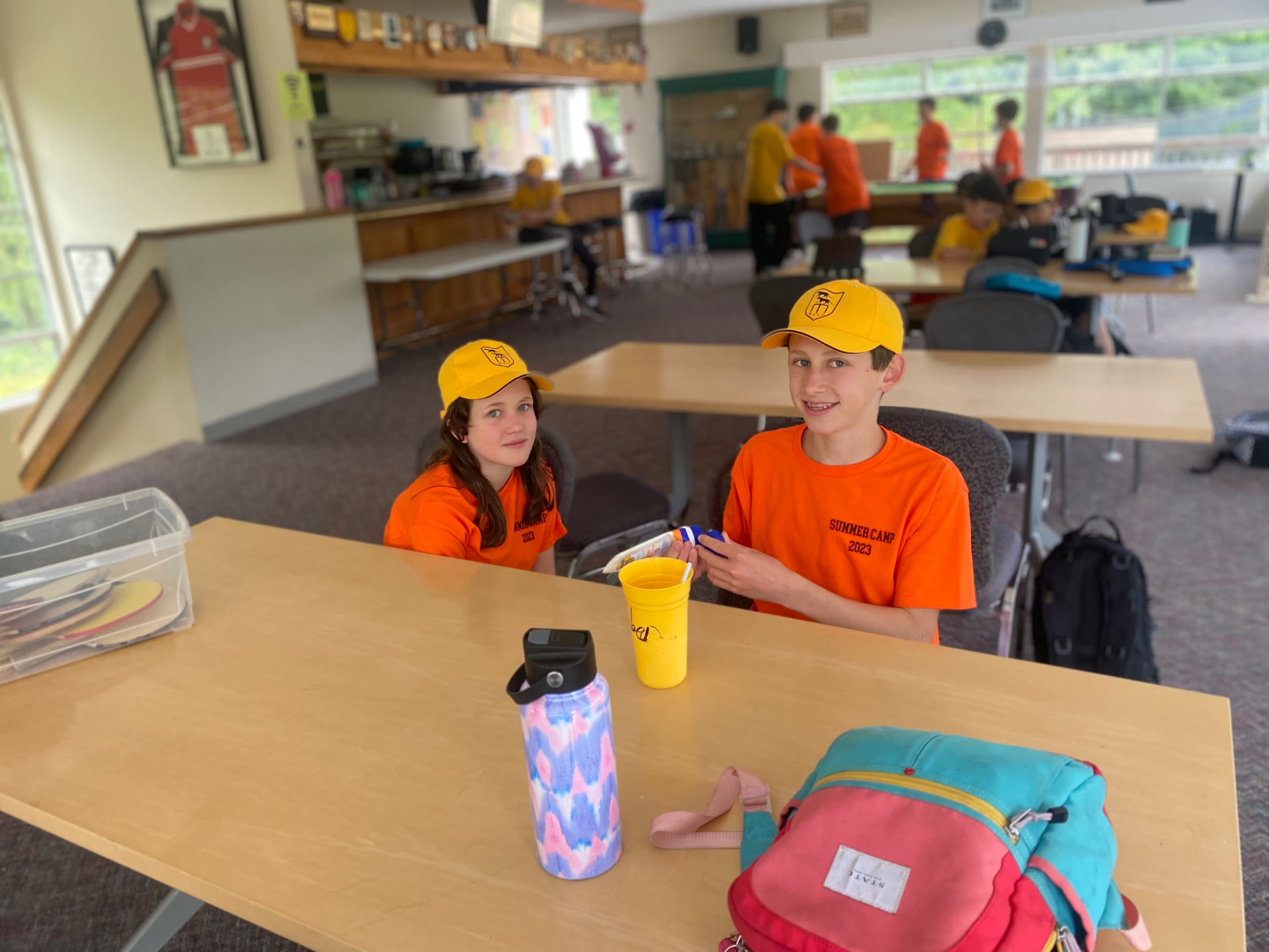 Two children wearing orange shirts and yellow hats sit at a long table in a room filled with other kids in the background.