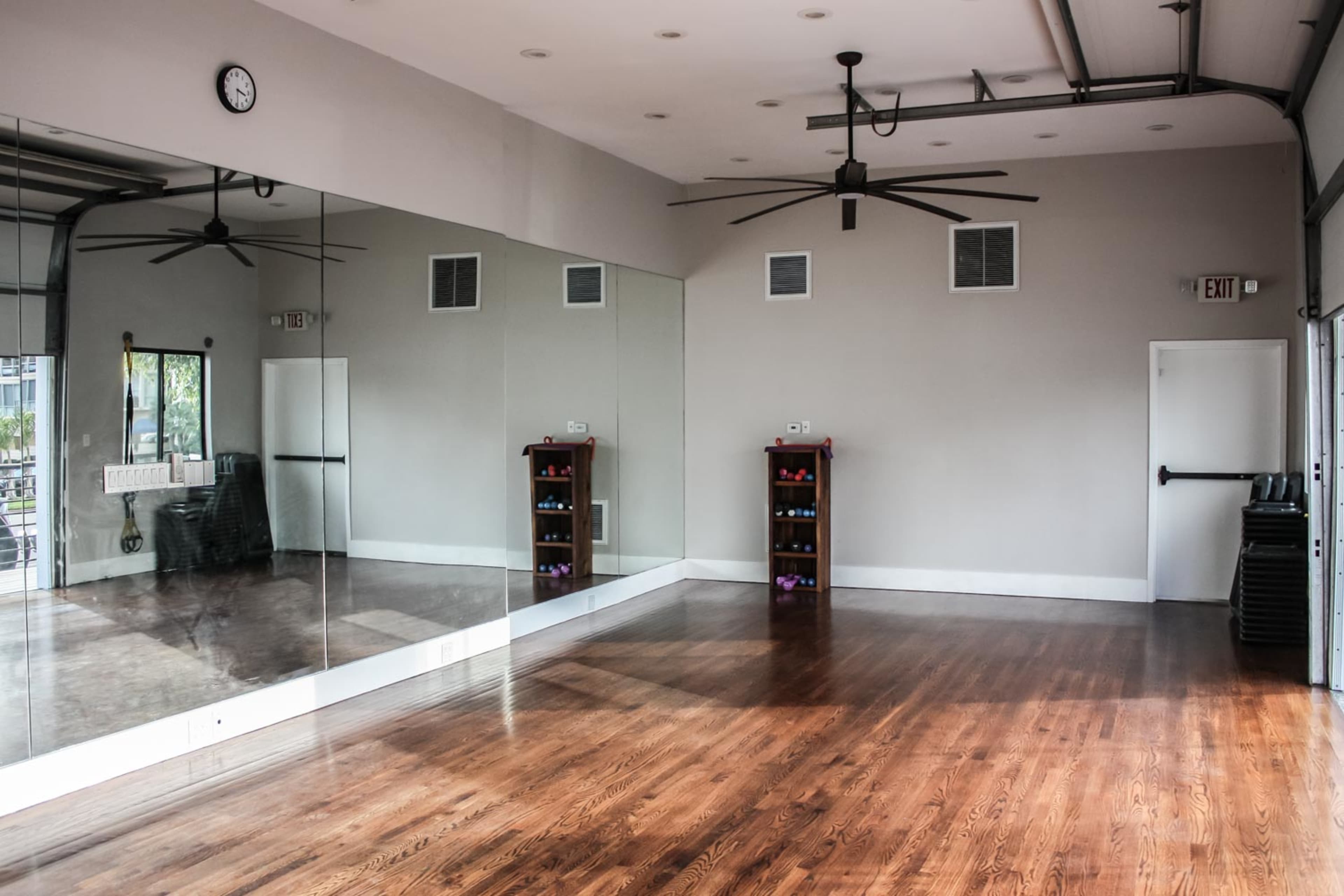 An empty exercise studio features wooden floors, large mirrors, wall-mounted fans, and organized shelves with weights.
