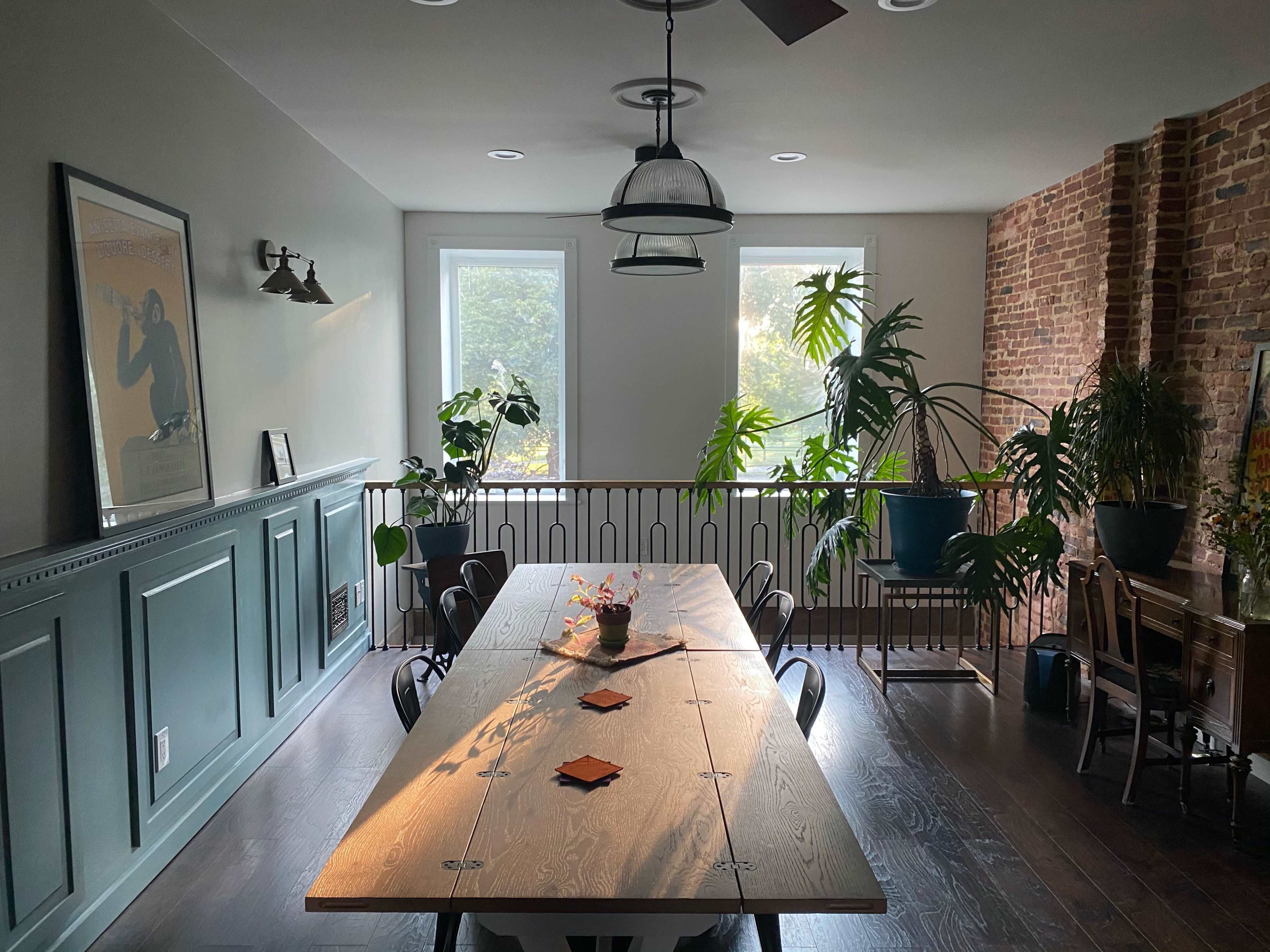 A spacious dining area features a long wooden table surrounded by chairs, with large windows and various indoor plants accentuating the brick and paneled walls.