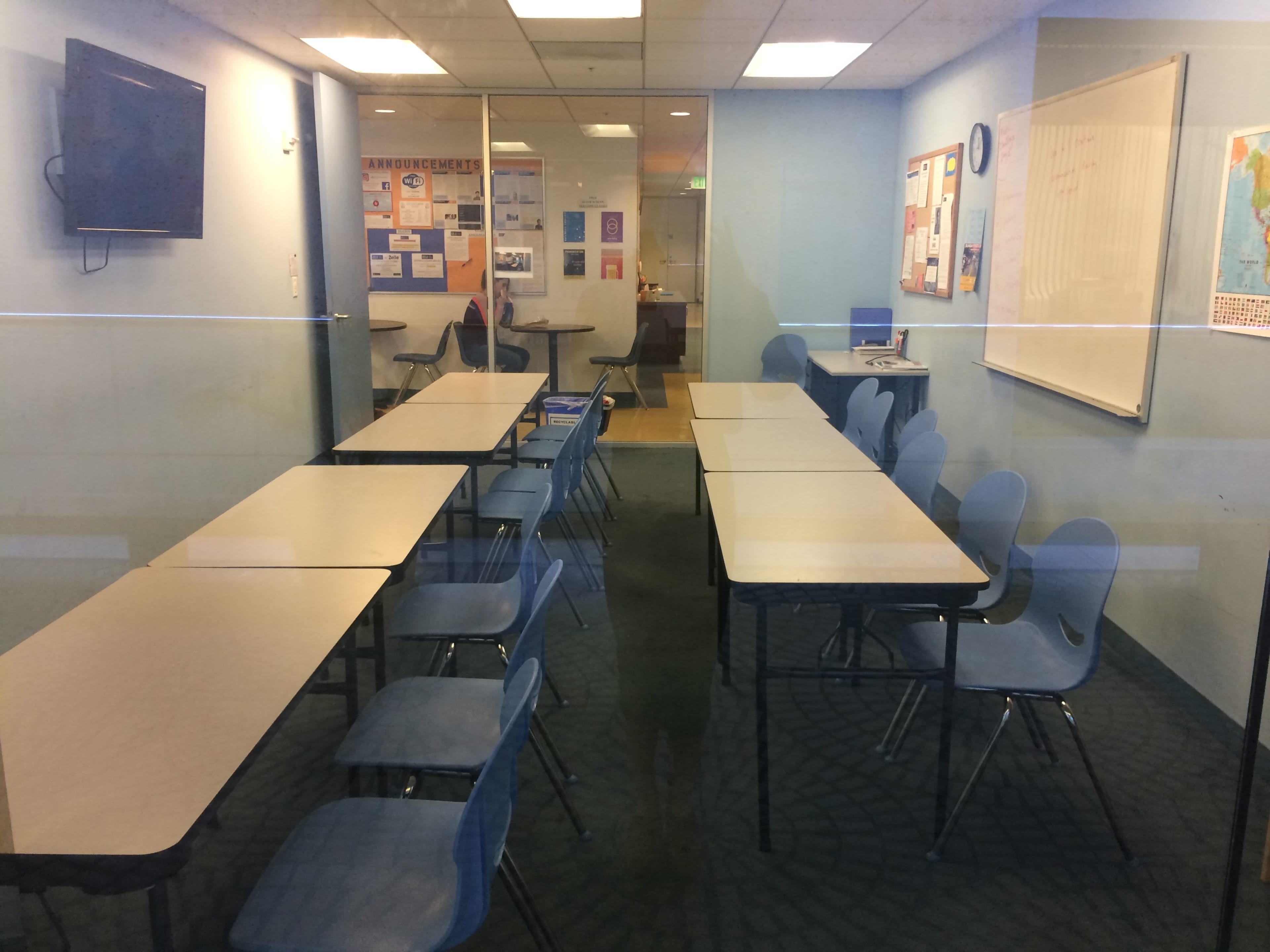 A classroom with several tables arranged in a U-shape and blue chairs, visible through glass walls, with educational materials on the walls.