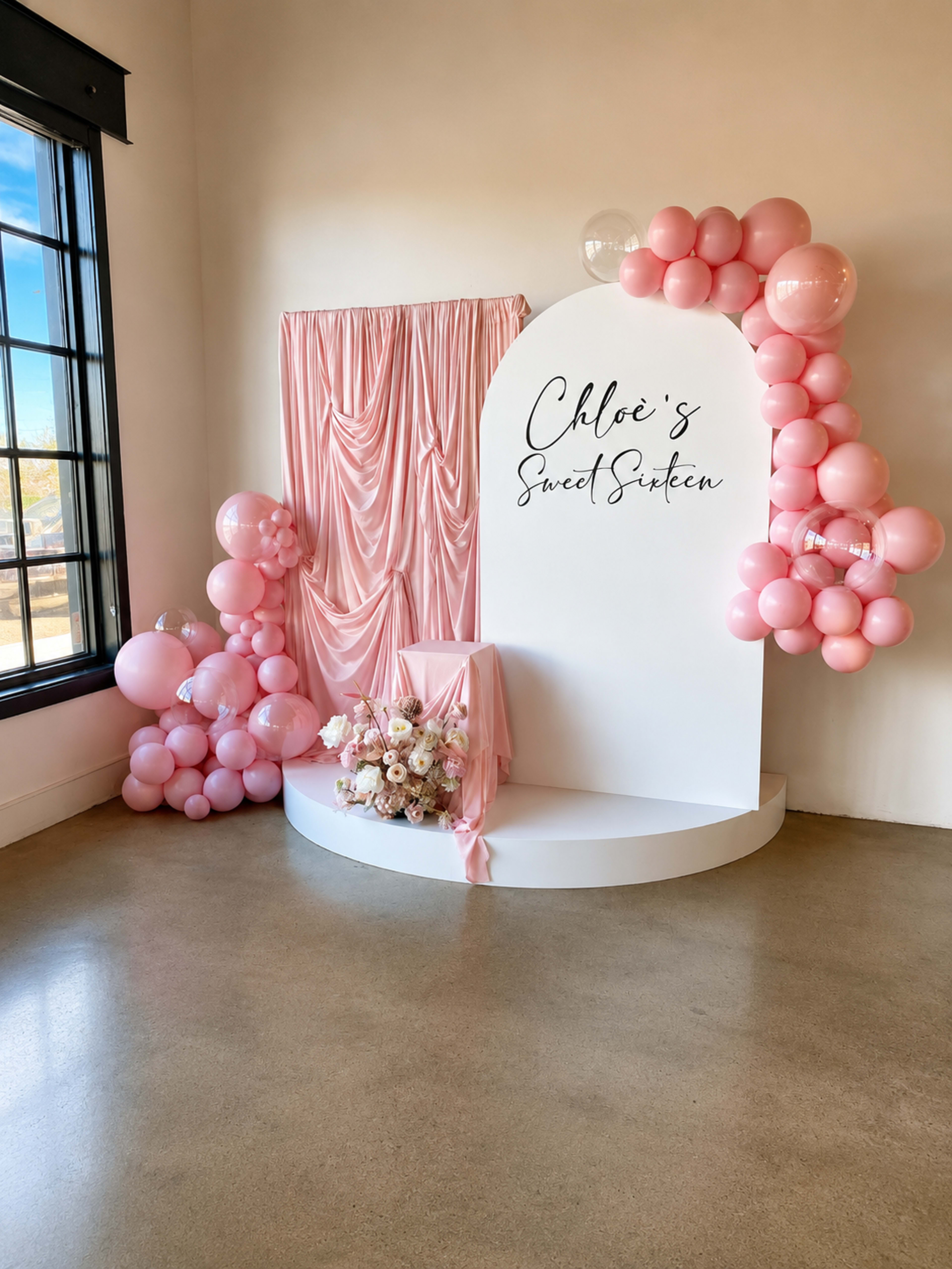The image shows a decorated event space featuring a large white backdrop with the text "Chloe's Sweet Sixteen," adorned with pink balloons and floral arrangements.