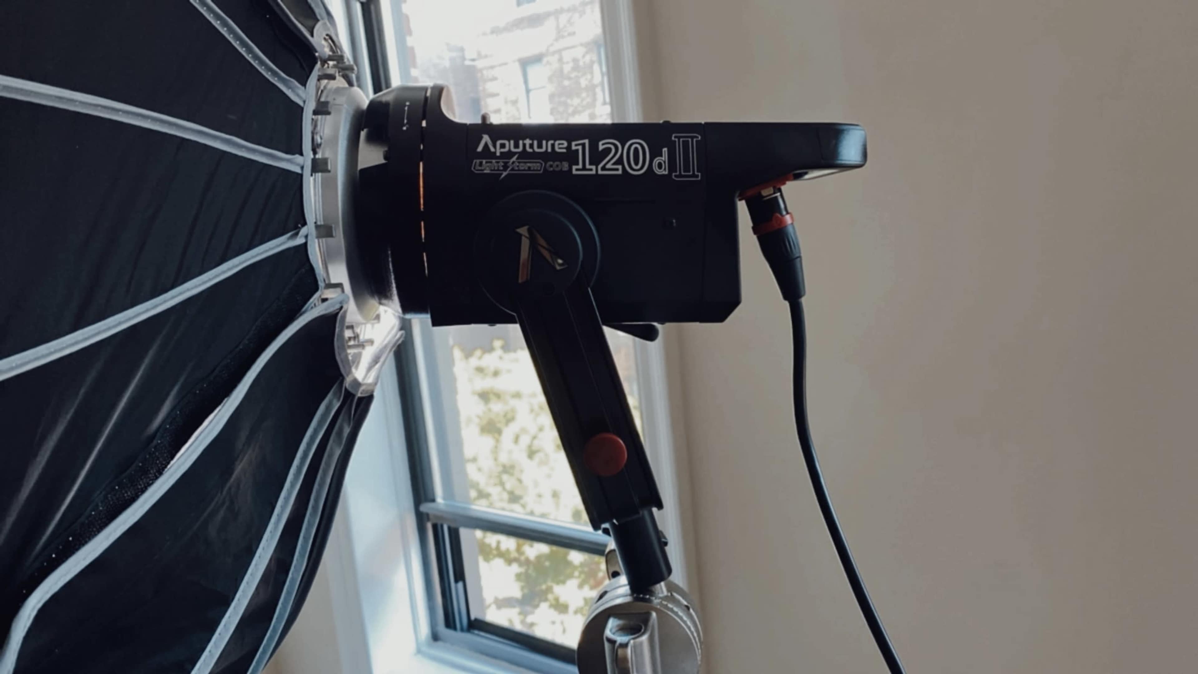 An Aputure 120d II lighting fixture mounted to a softbox, with a cable connected to it, placed near a window.