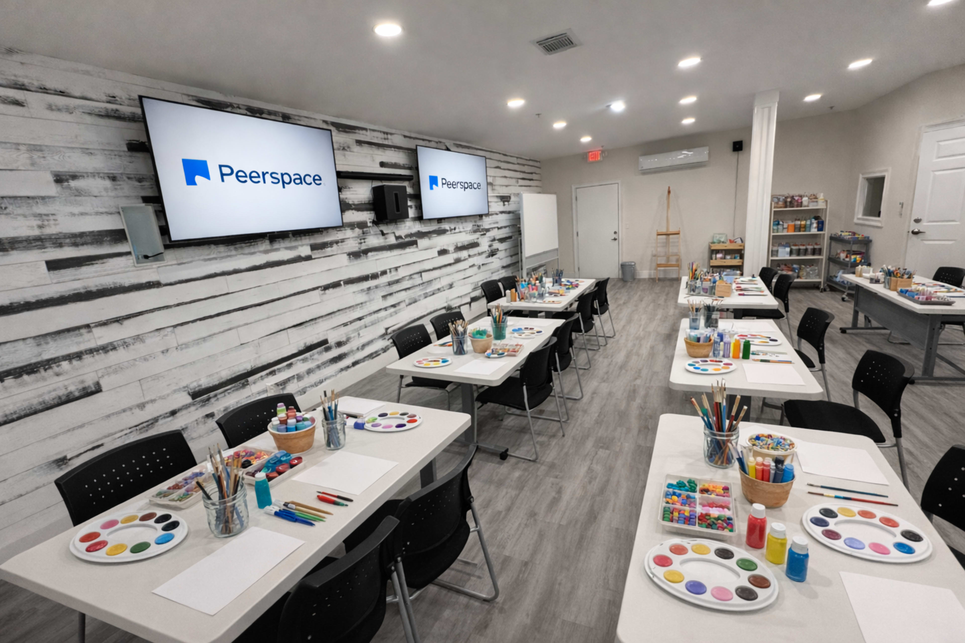 The image shows a well-organized art studio with multiple tables set up, each equipped with art supplies including paint, brushes, and paper, and two screens displaying the Peerspace logo.