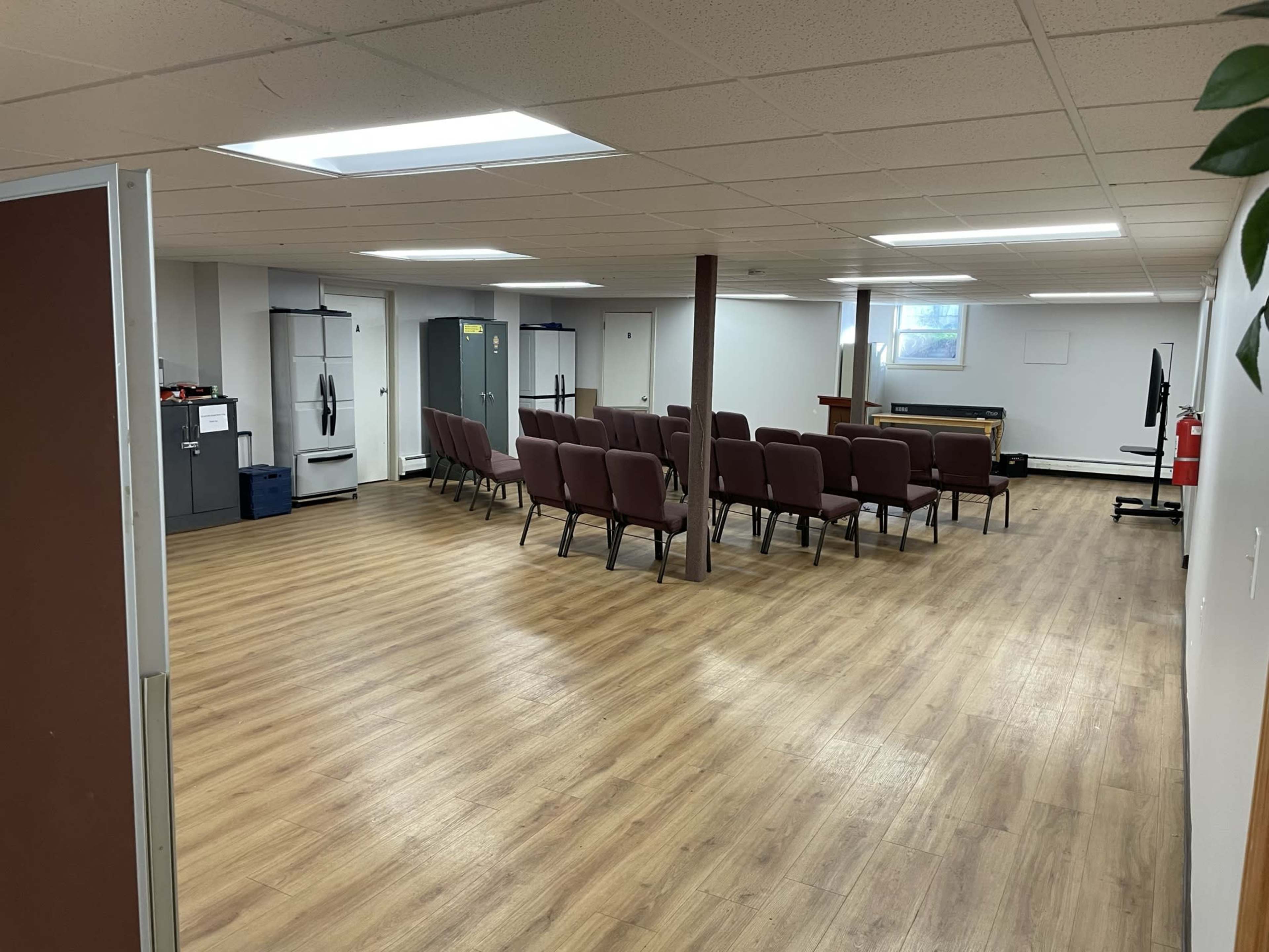 A spacious, well-lit room with rows of chairs facing a small stage or podium, along with various appliances and a television in the background.