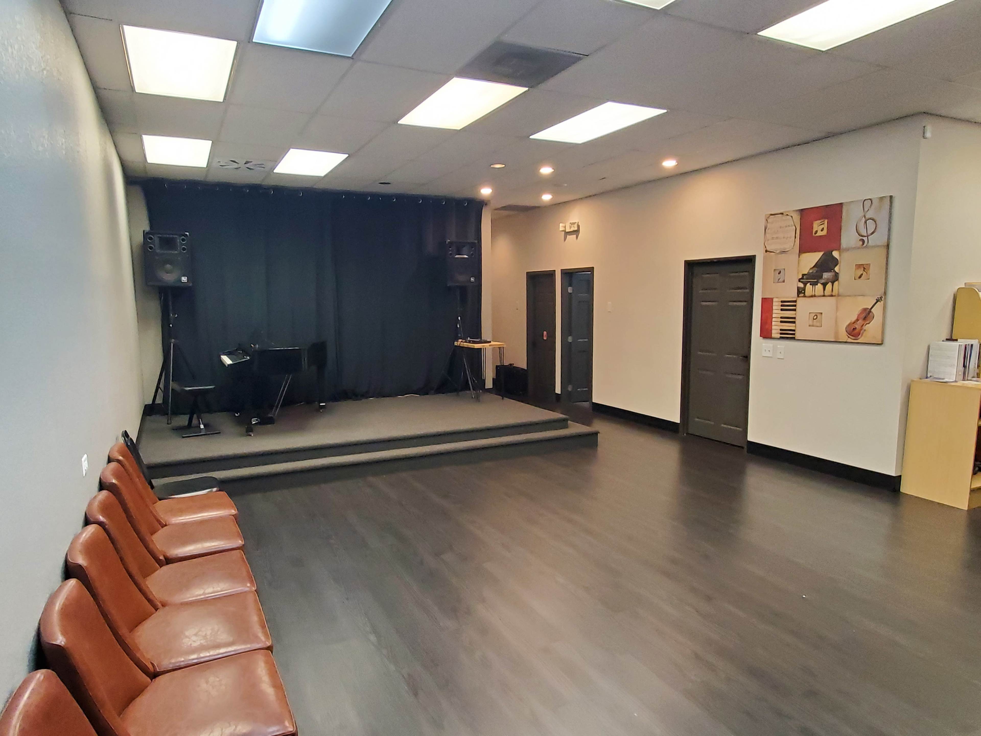 The image shows a minimalist event space with a small stage, a piano, and several rows of brown chairs arranged in front of it.