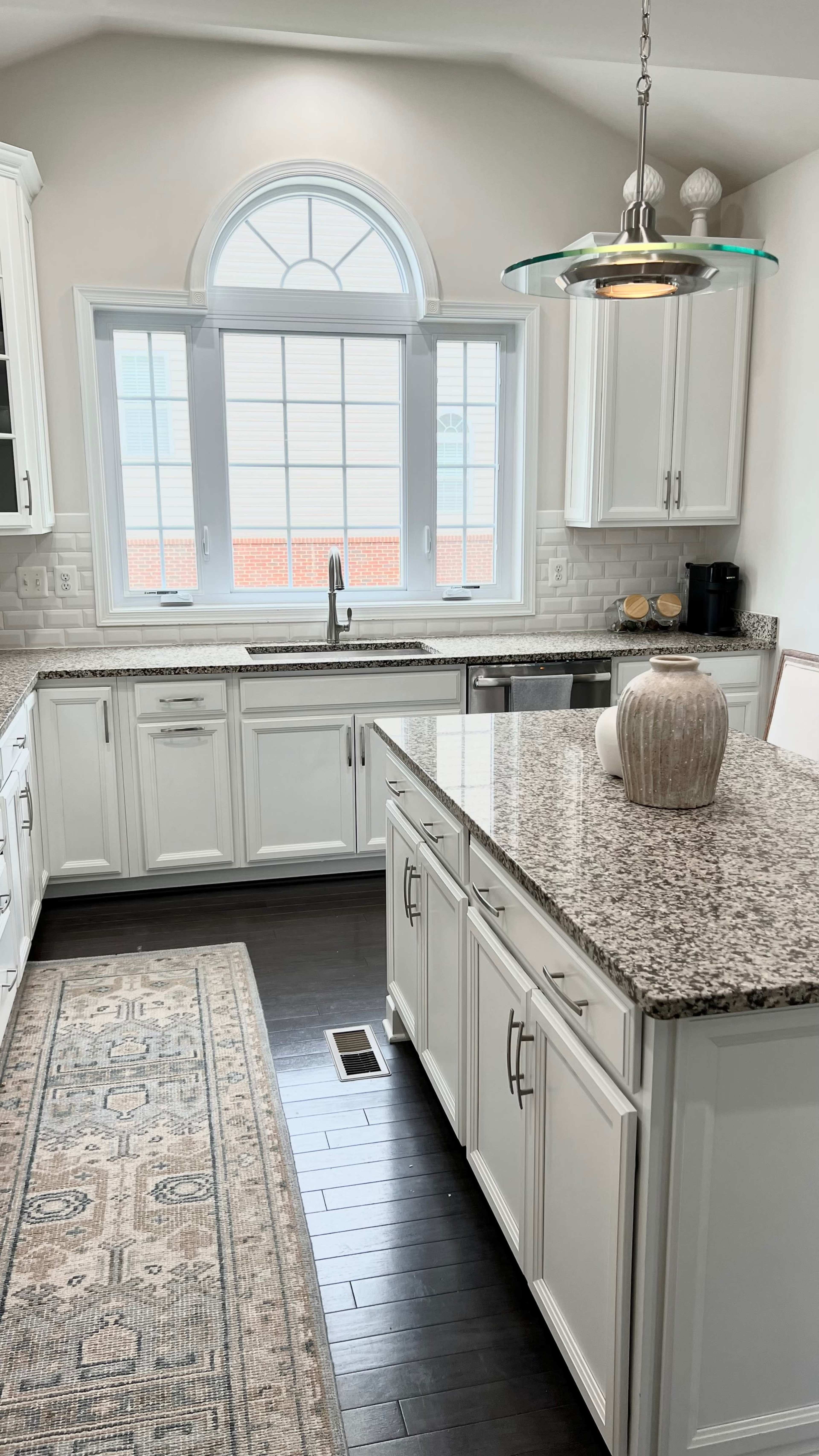 A modern kitchen features white cabinets, a granite countertop, and a large window above the sink that allows natural light to fill the space.