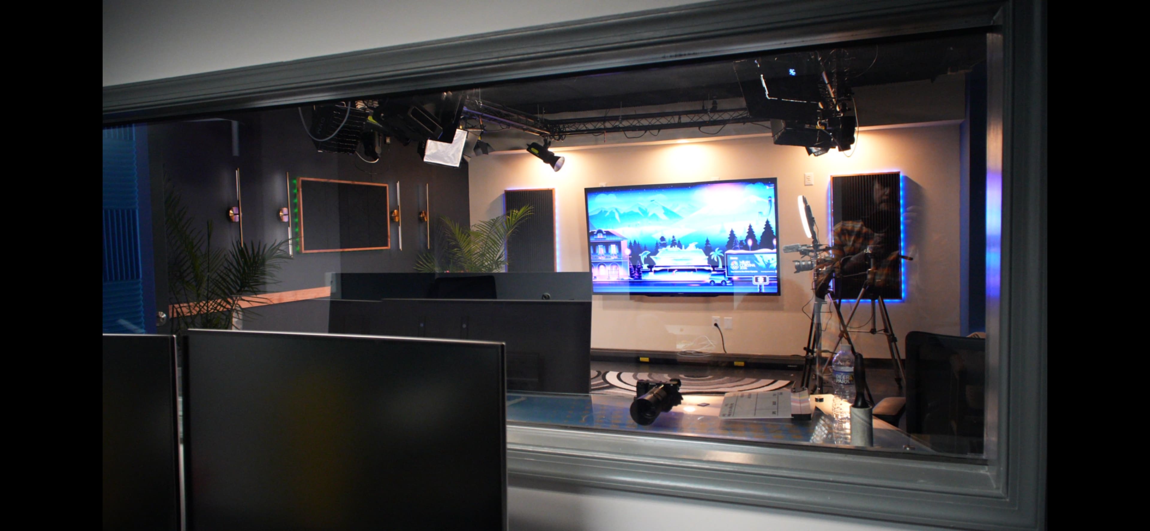 A view of a studio space through a glass window, featuring a large screen displaying a landscape and various studio equipment in the foreground.