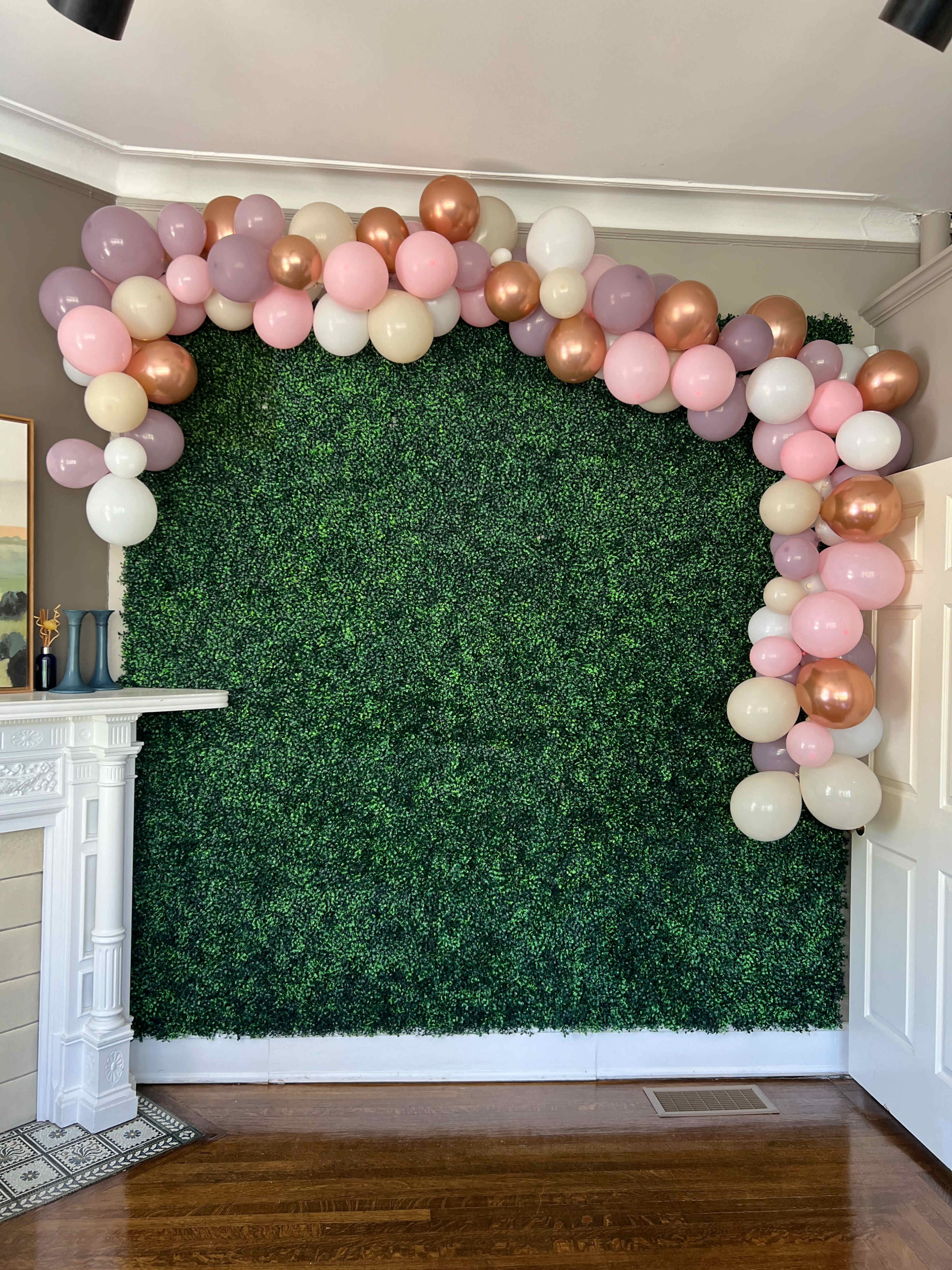 A decorated wall covered in artificial greenery, with a cluster of balloons in various colors arranged at the top.