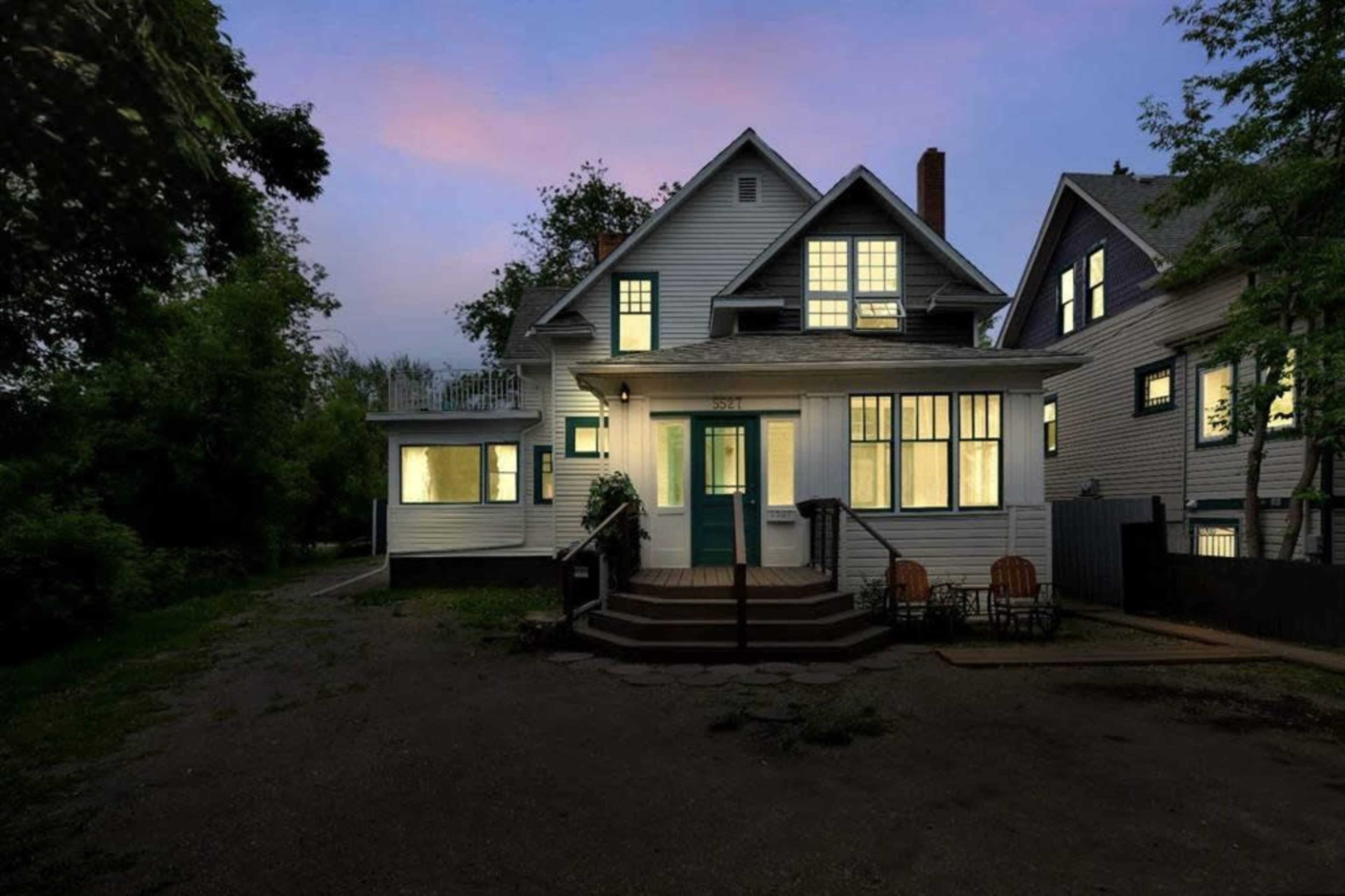 A two-story house with a manicured front yard and several large windows is illuminated at dusk.
