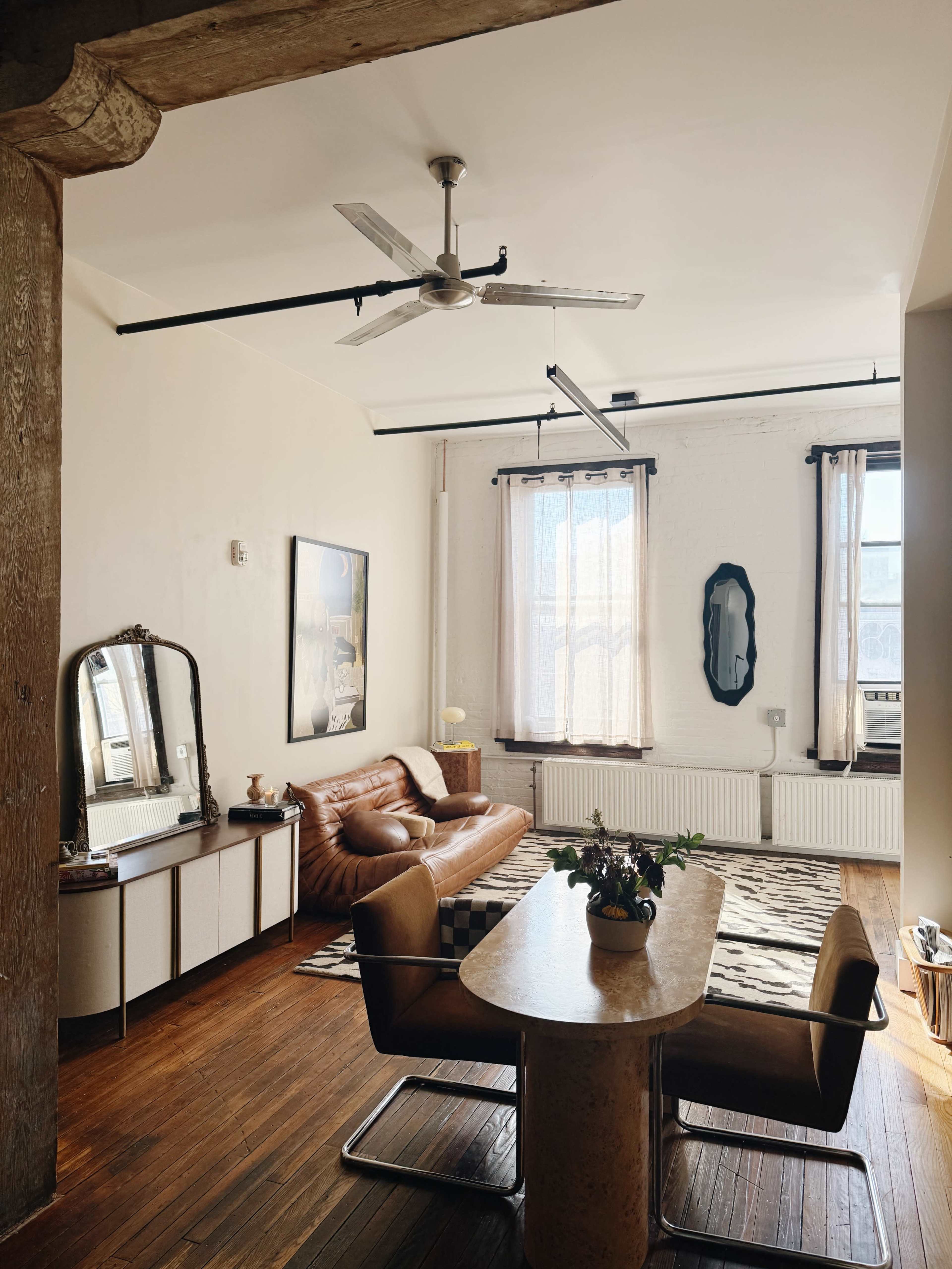 The image shows a minimalist living room featuring a brown leather sofa, an oval mirror, a dining table, and large windows with sheer curtains.