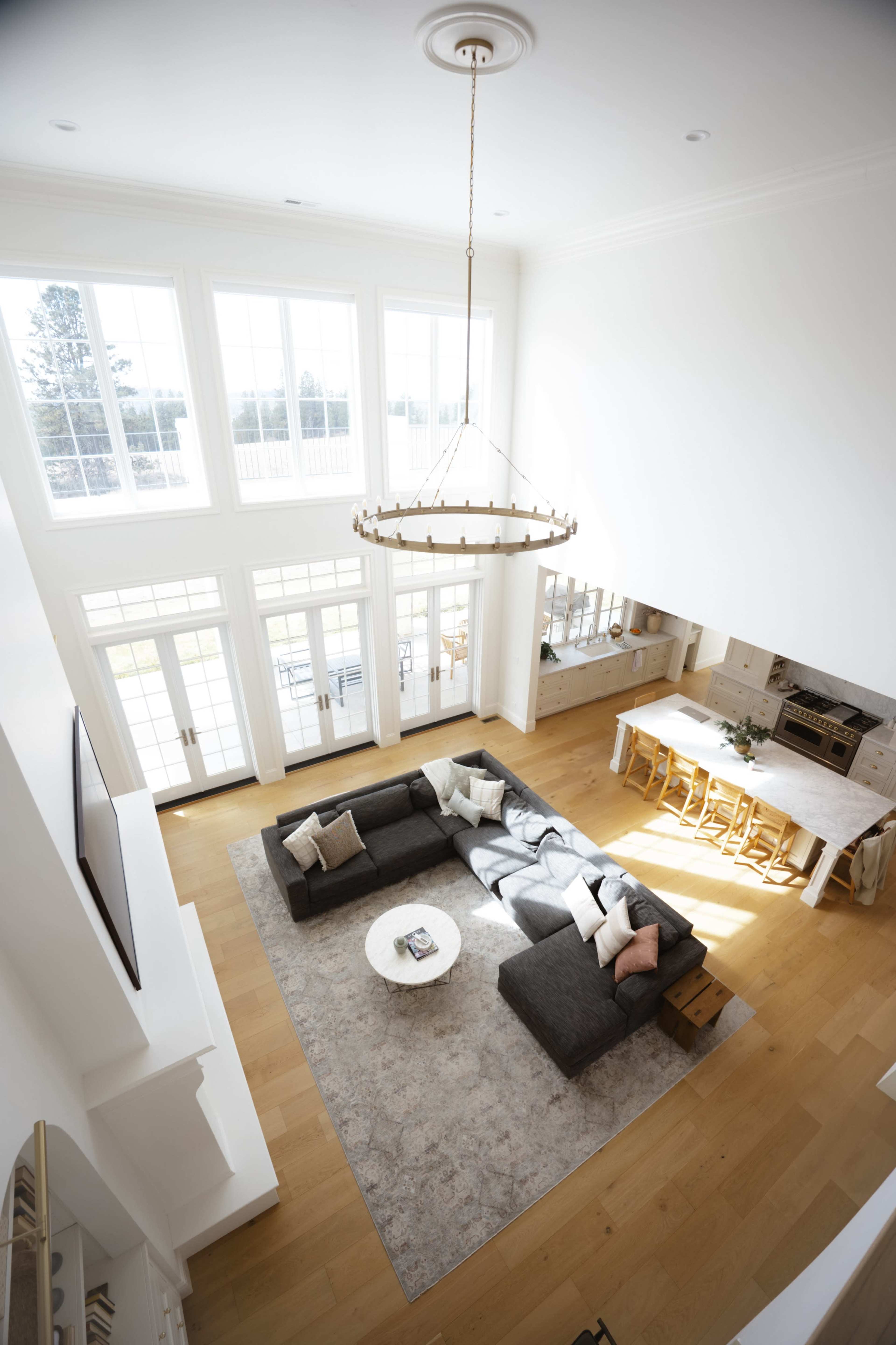 A spacious, modern living room features a large sectional sofa and a round coffee table, illuminated by natural light from tall windows and accented by a chandelier above.