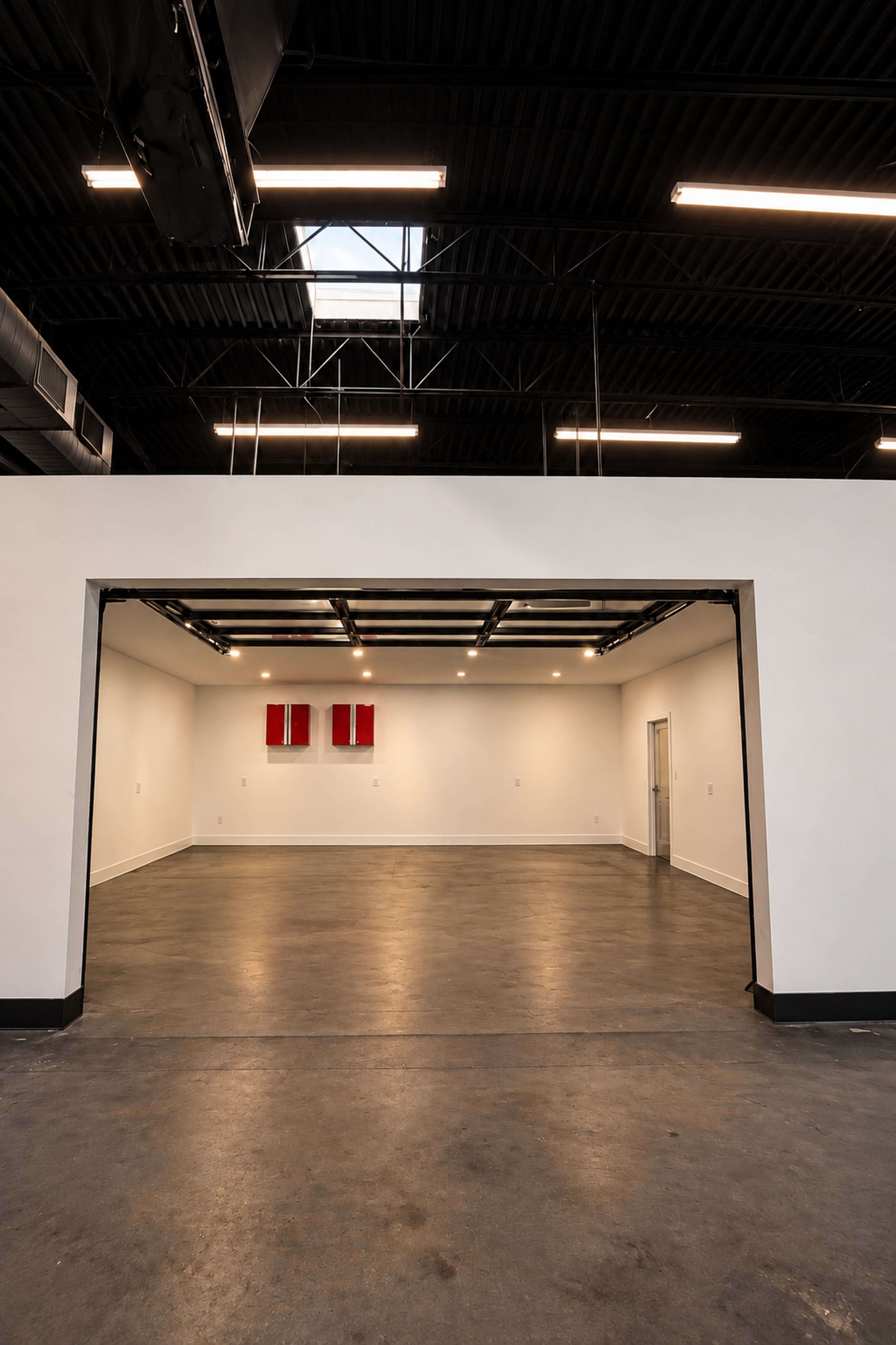 The image shows a spacious, empty room with a high ceiling, featuring a large opening that leads to another area, illuminated by overhead lights.