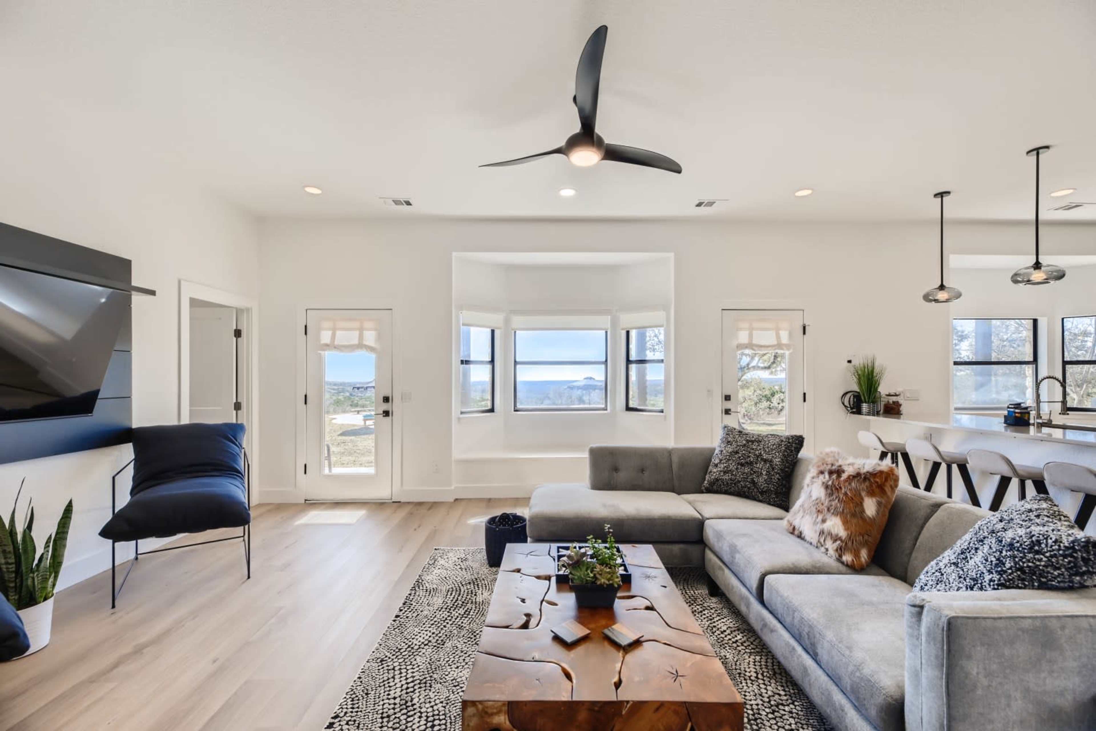 The living room features a large sectional sofa, a wooden coffee table, and a wall-mounted TV, with large windows that provide a view of the outdoors.