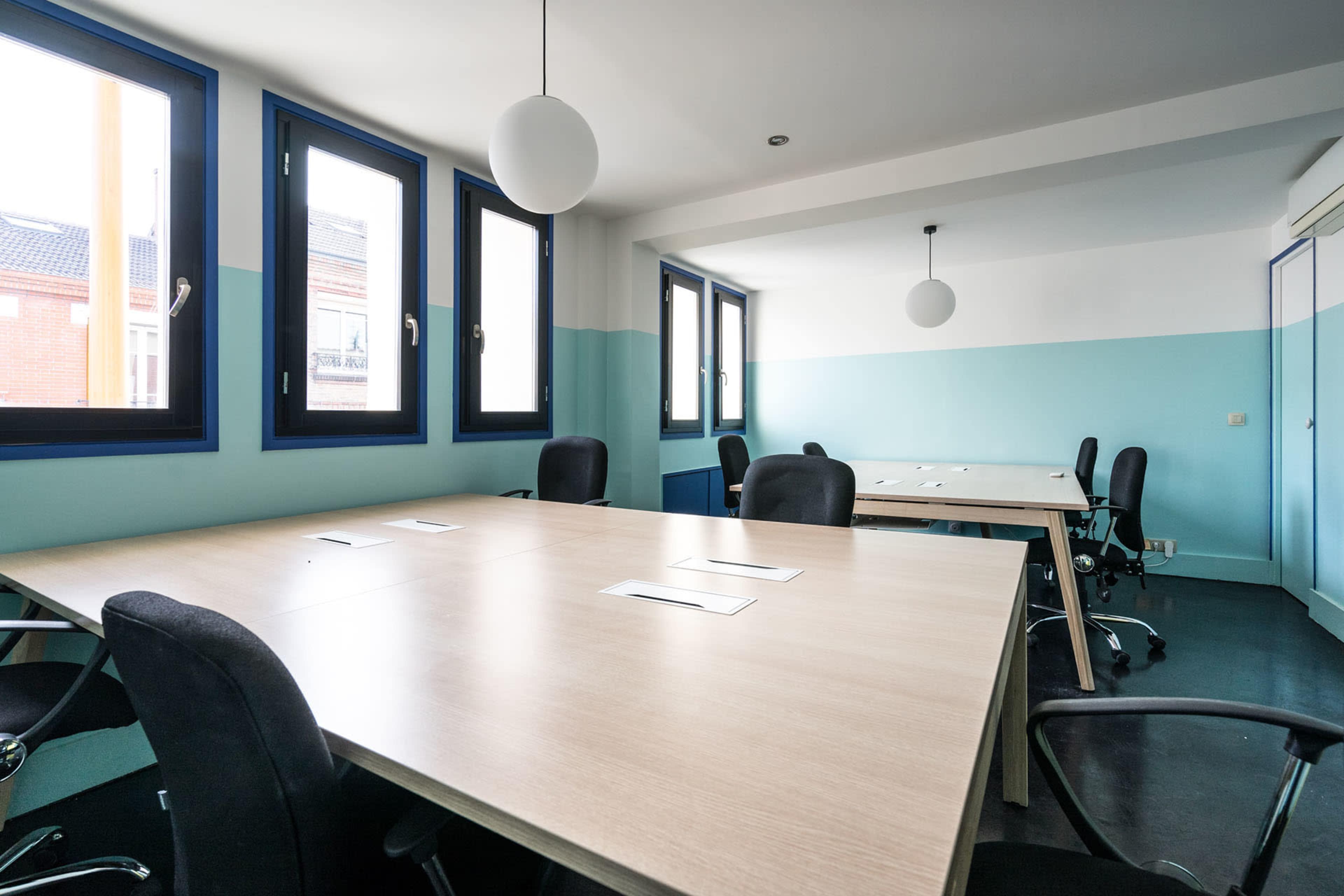 A modern conference room features several large tables with ergonomic chairs, blue and white walls, and multiple windows allowing natural light.