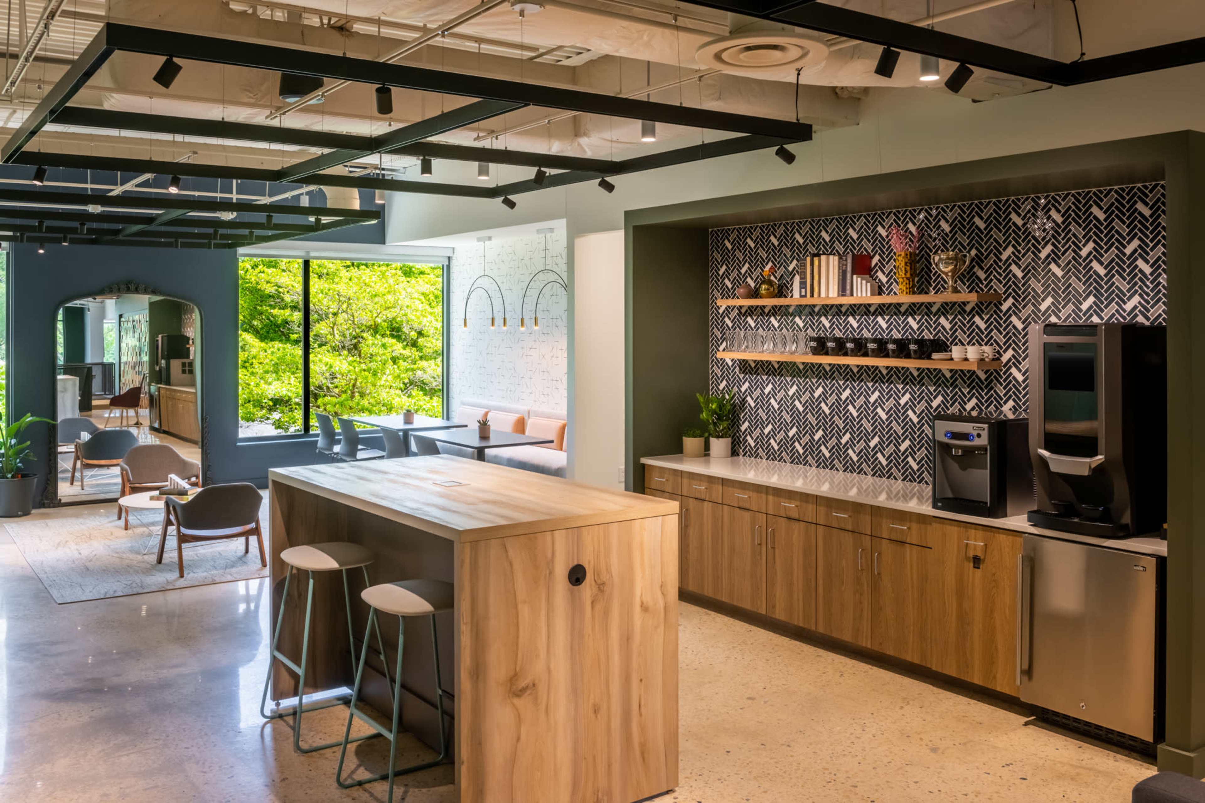 The image shows a modern kitchen and dining area with a wooden island, a coffee machine, and seating alongside large windows overlooking greenery.