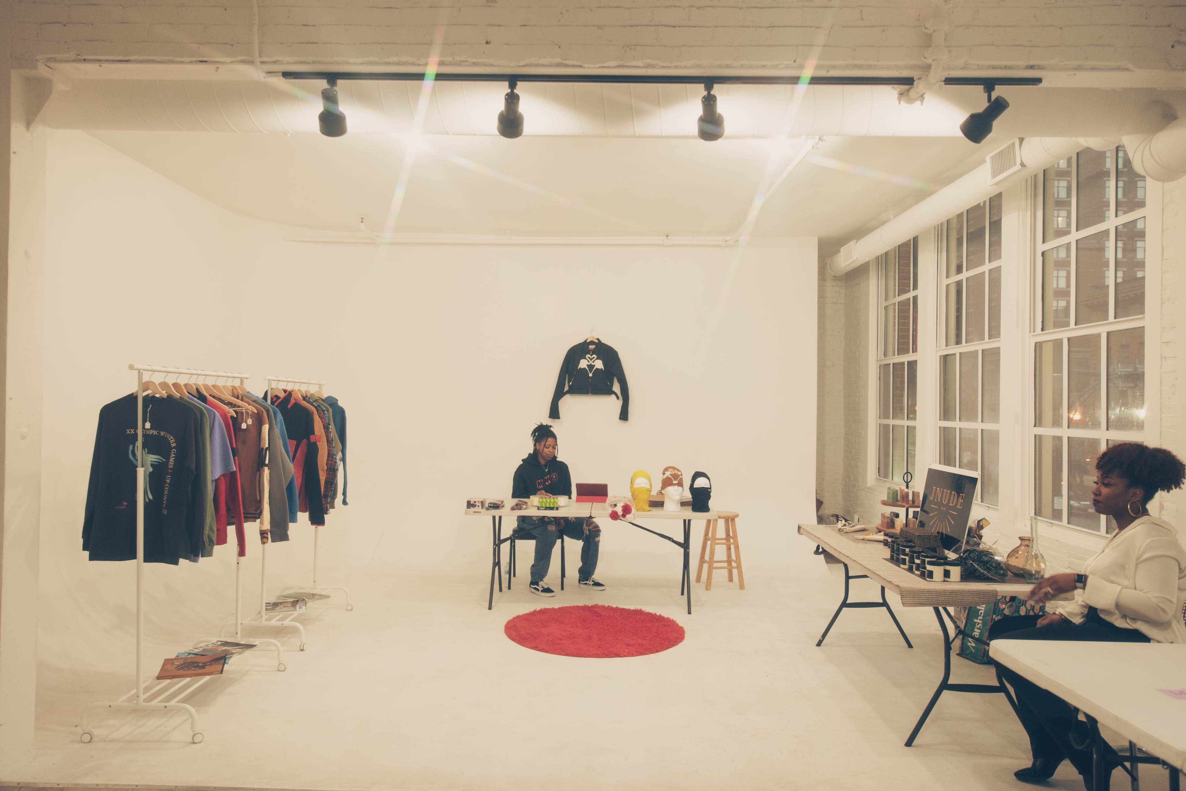 A person sits at a table in a spacious, well-lit gallery featuring clothing on racks and various items displayed on tables.