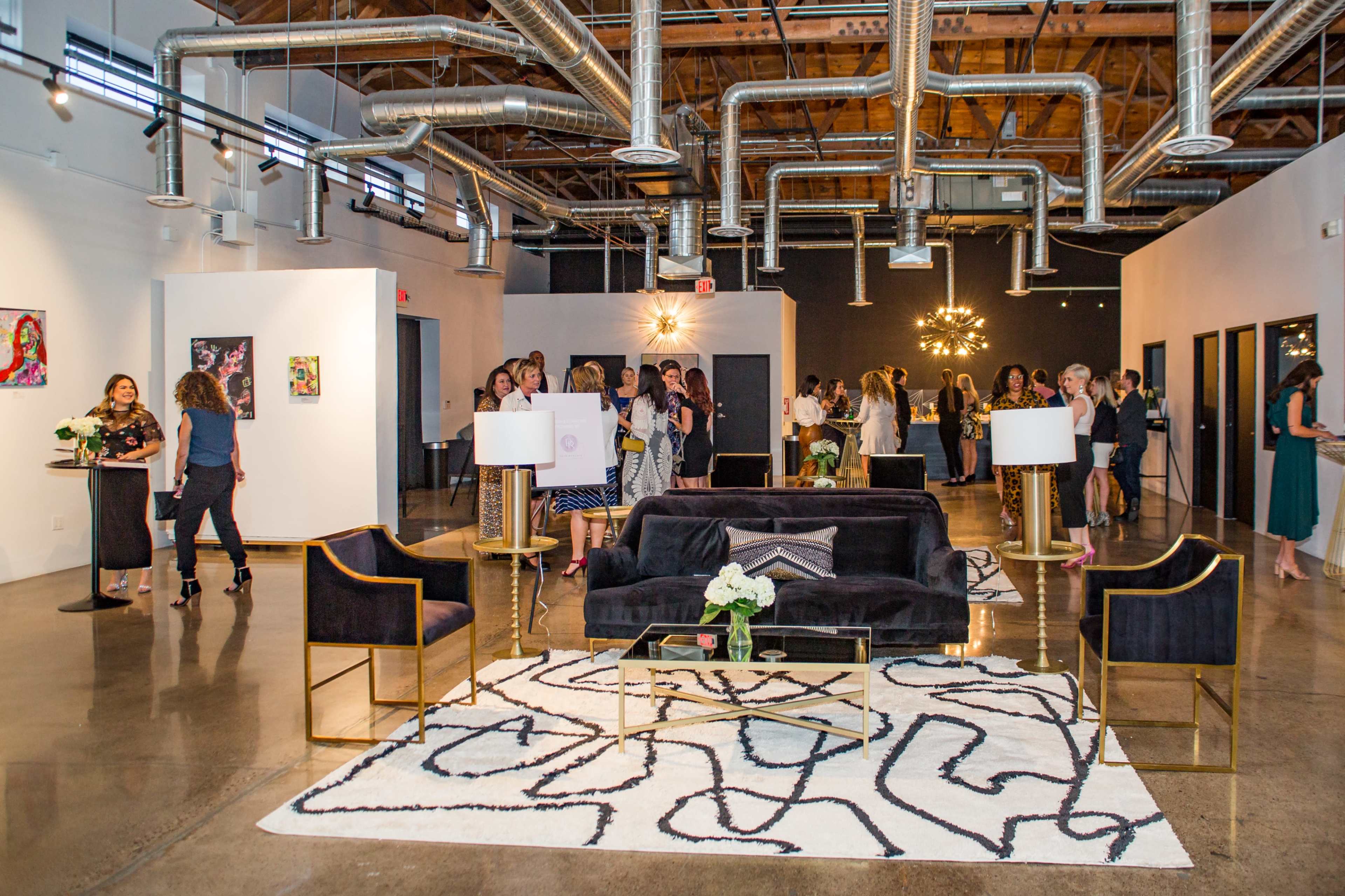 A modern event space with guests mingling, featuring a seating area with a black sofa, decorative lamps, and artwork displayed on the walls.
