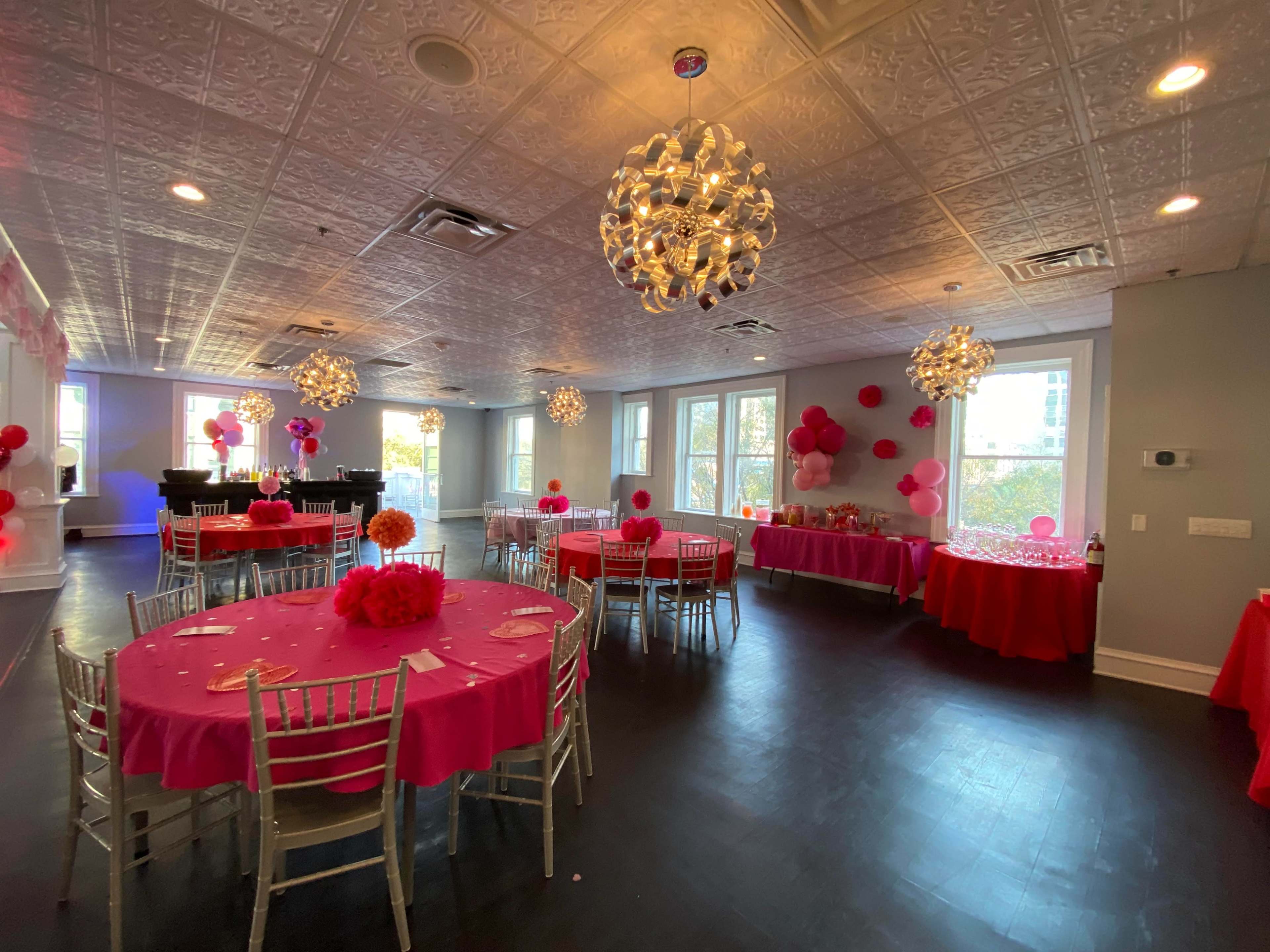 A spacious event room features tables decorated with pink tablecloths and centerpieces, illuminated by decorative chandeliers.