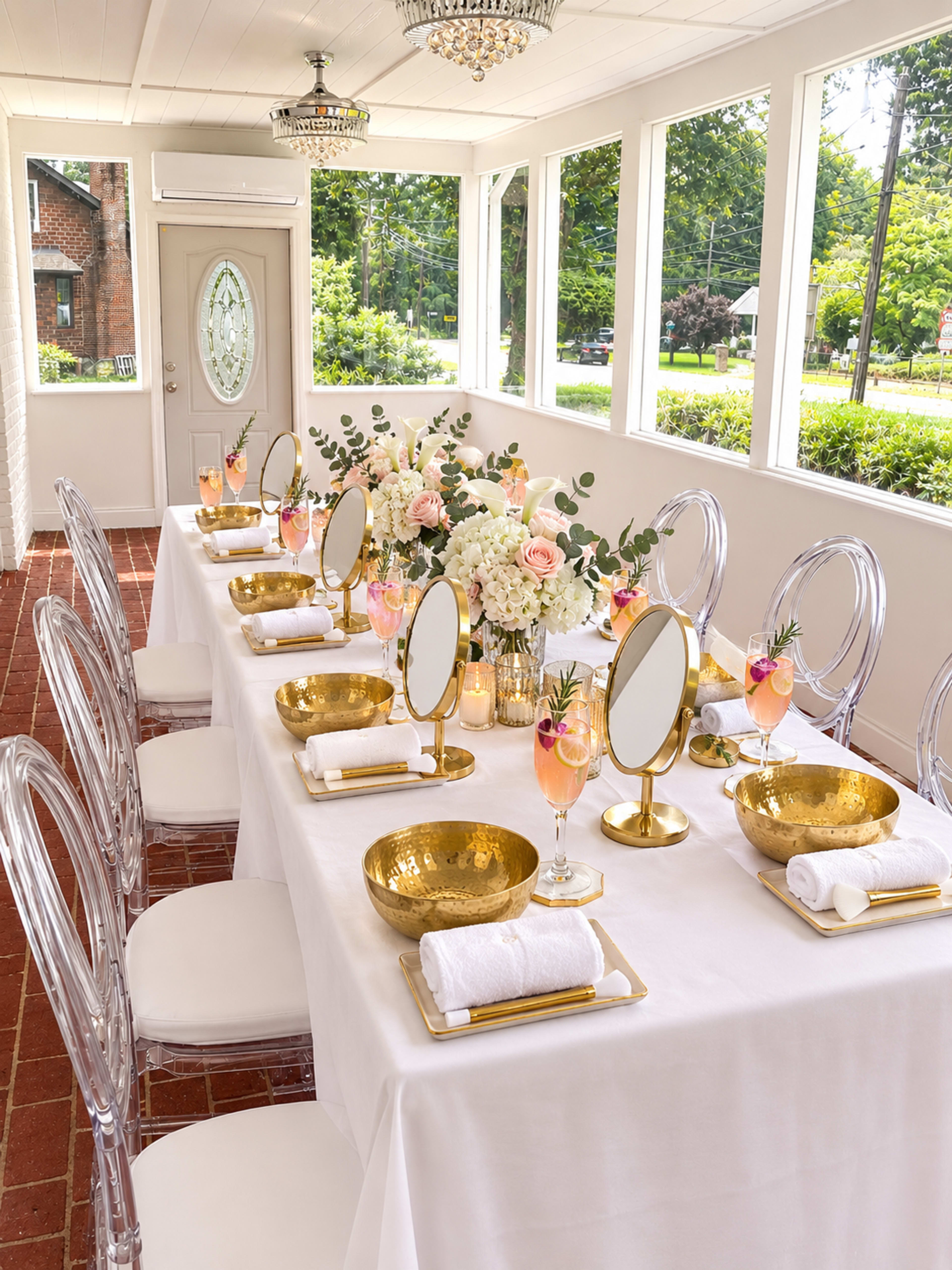 A long, elegantly arranged dining table is set with gold-accented bowls, mirrors, and floral centerpieces in a bright, sunlit room.