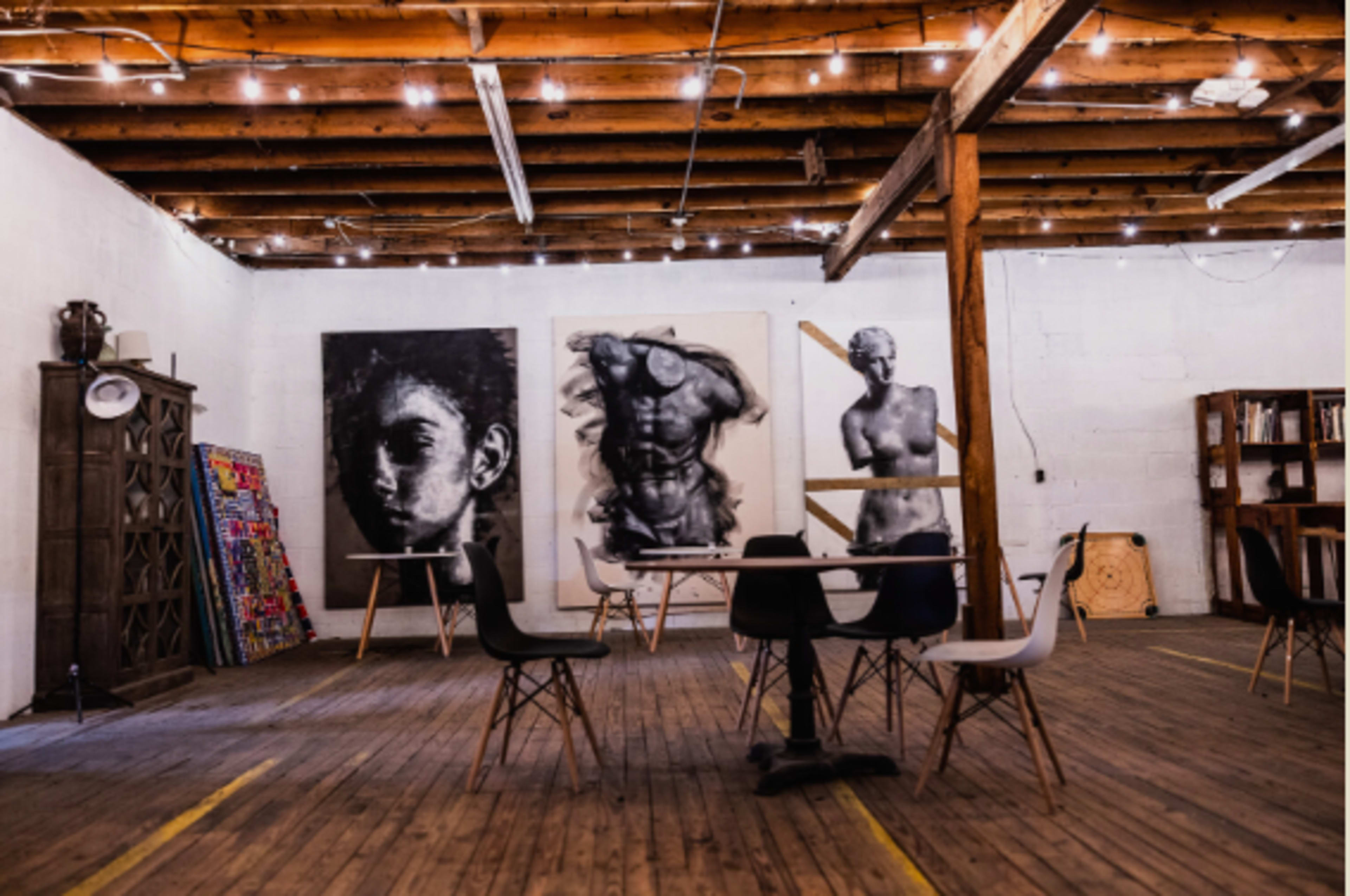 An art studio with wooden floors, featuring several large artworks on the walls, scattered chairs, and warm string lights overhead.