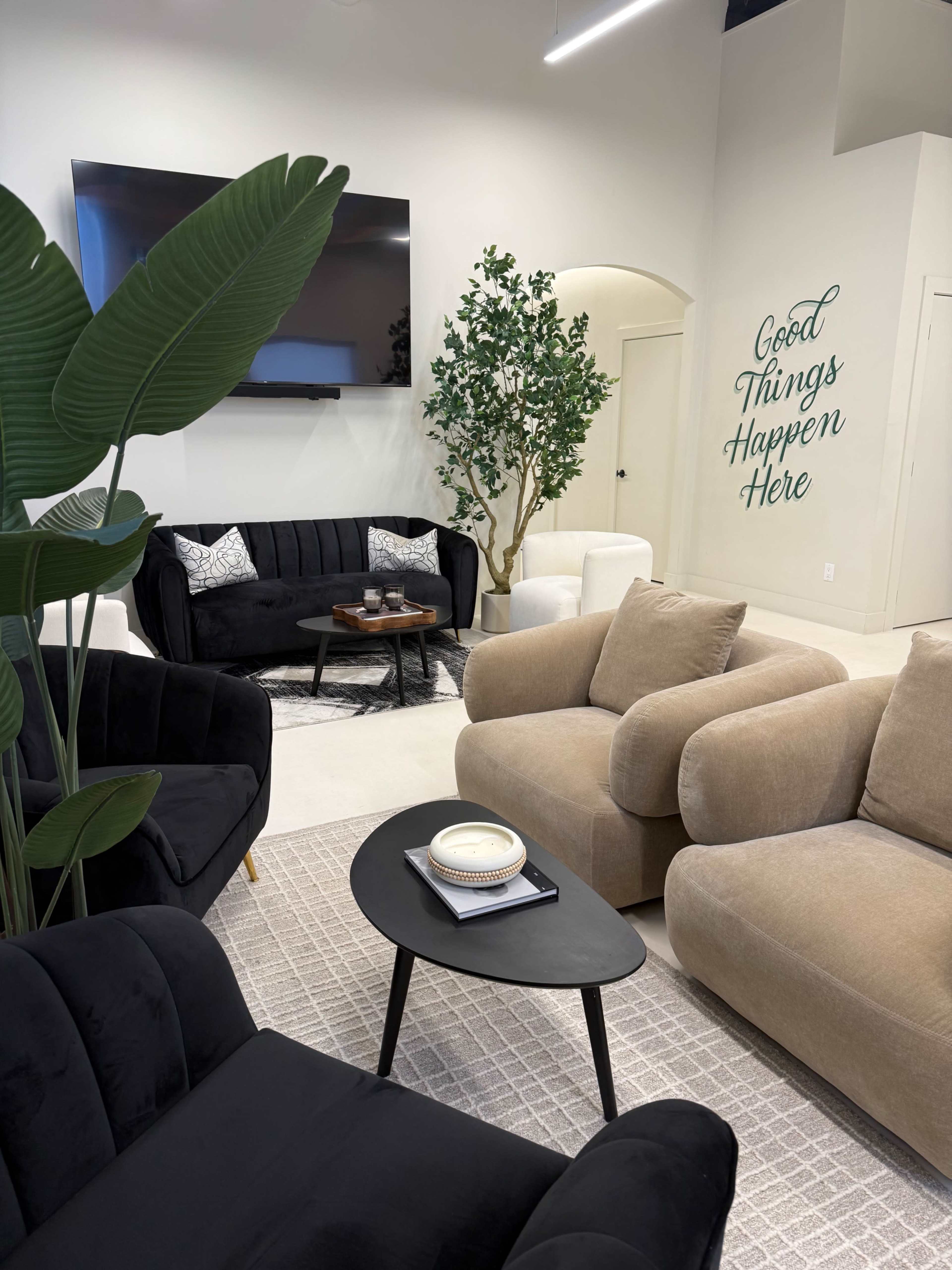 A modern lounge area features black and beige sofas, a circular black coffee table, lush greenery, and a wall with the phrase "Good Things Happen Here."