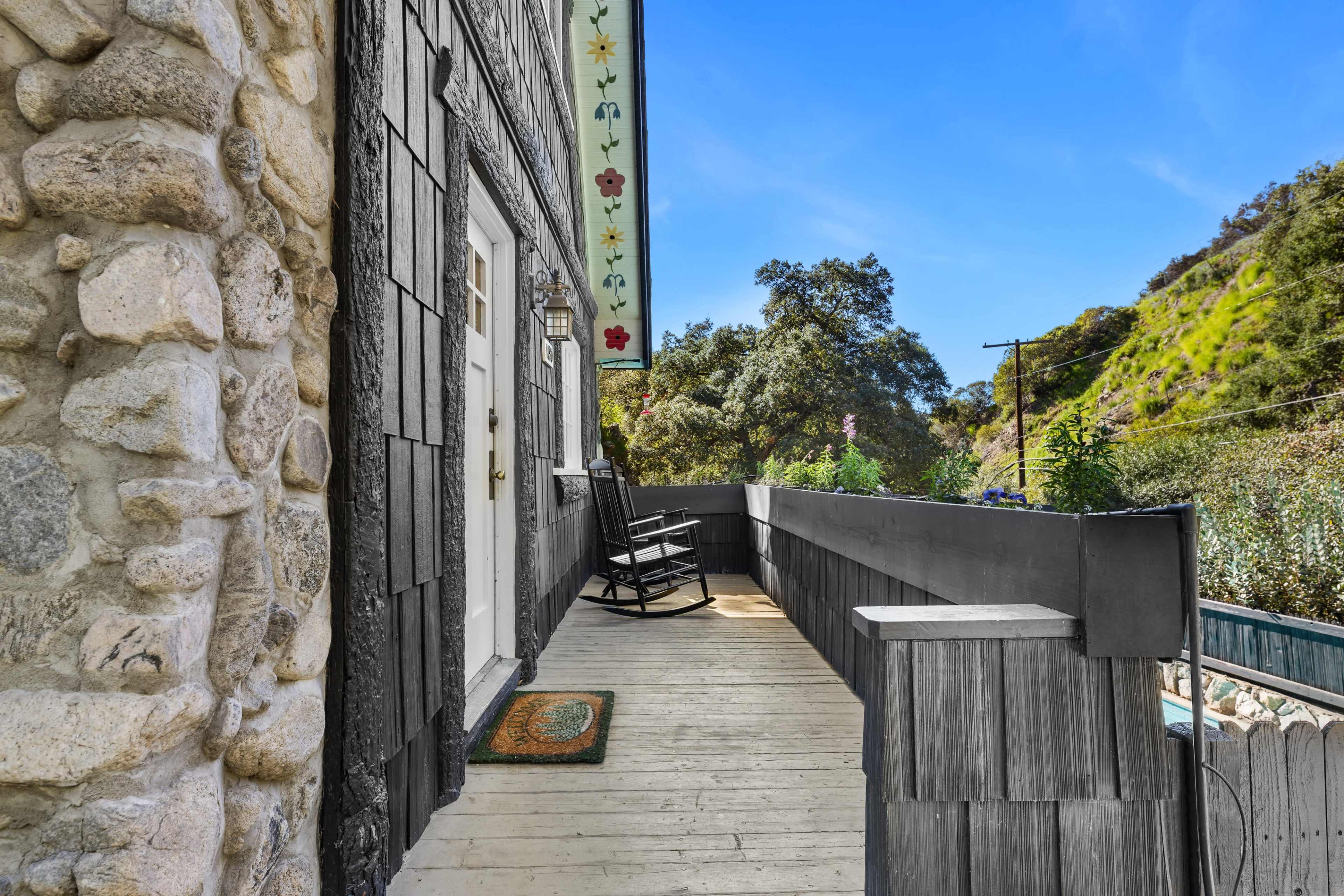 The image shows a wooden porch with a rocking chair and a stone wall, surrounded by greenery and hills in the background.