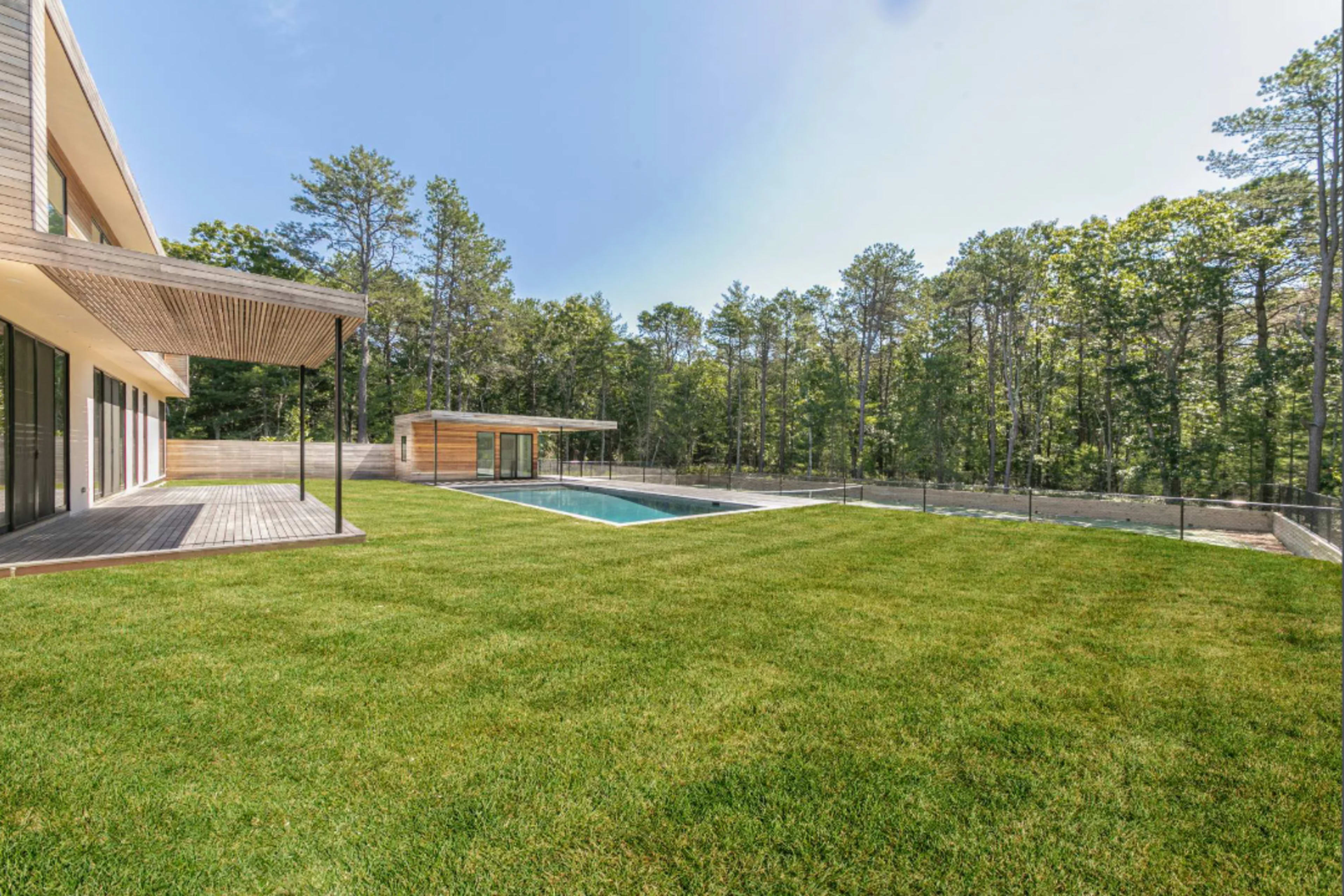 A modern house with large windows overlooks a rectangular swimming pool surrounded by a neatly manicured lawn and forested area.