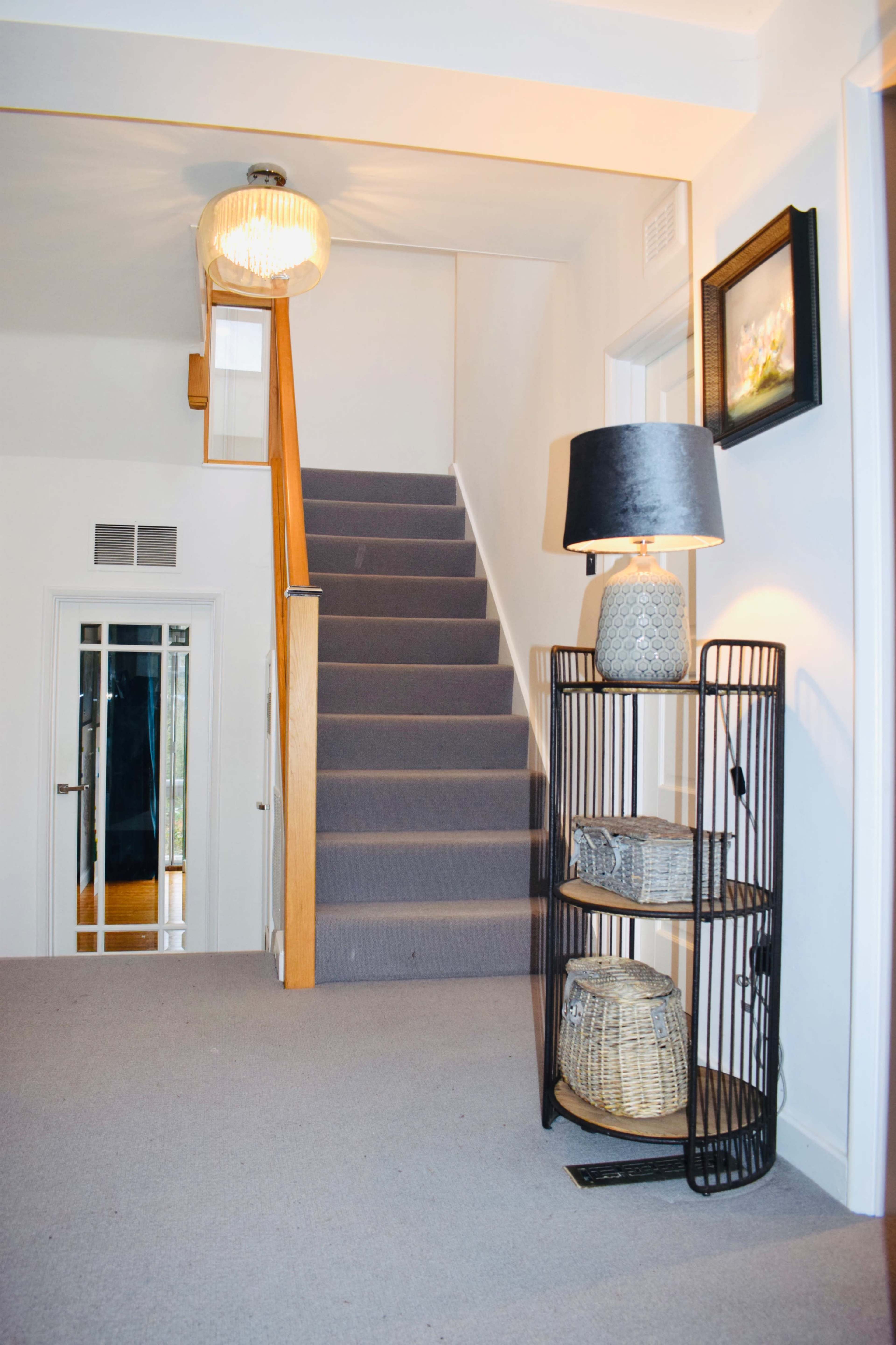 A well-lit hallway with a staircase leading to an upper floor, a lamp on a tiered stand, and a framed picture on the wall.