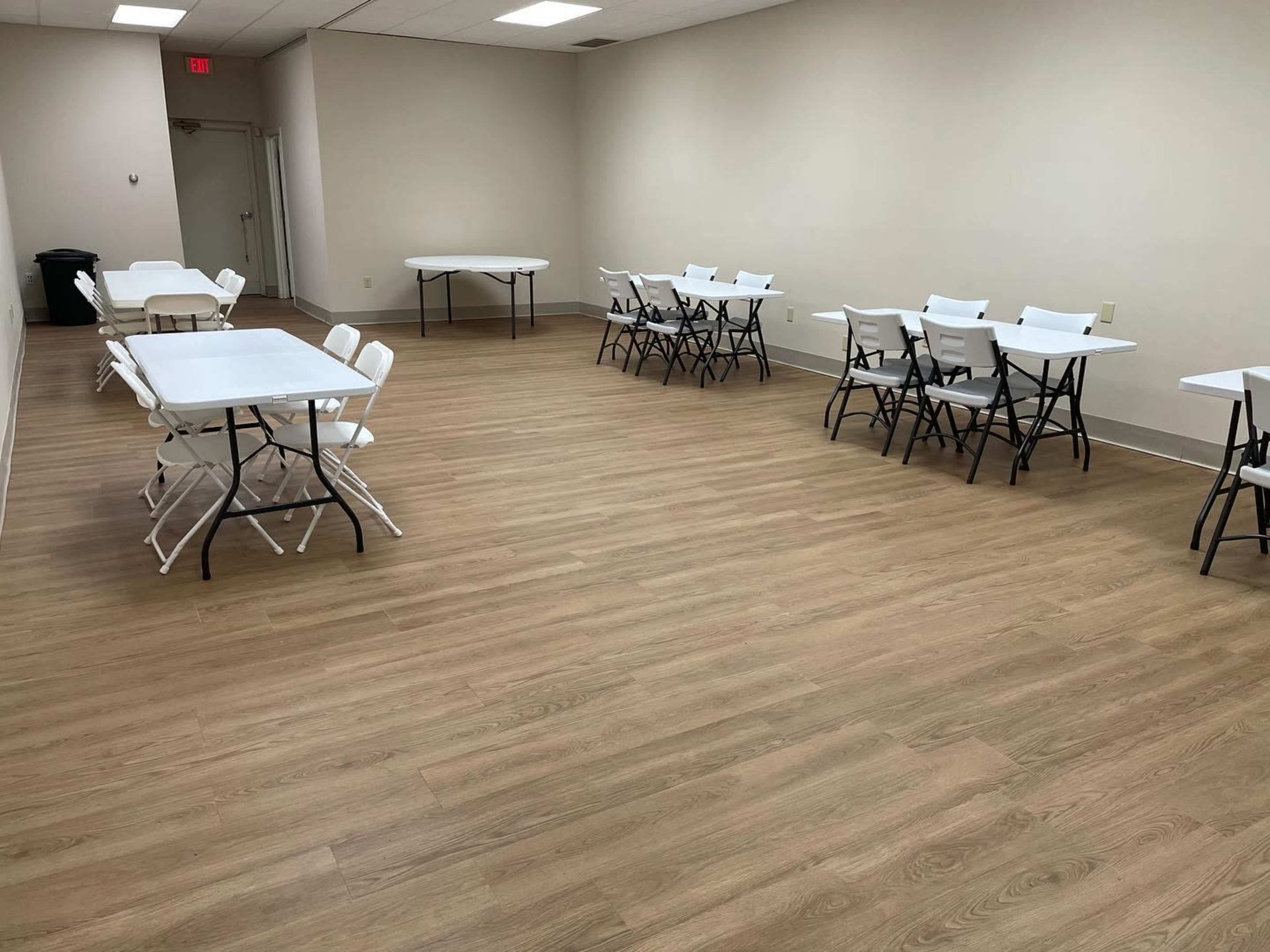 A bright, empty room with several white folding tables and chairs arranged neatly on a wooden floor.