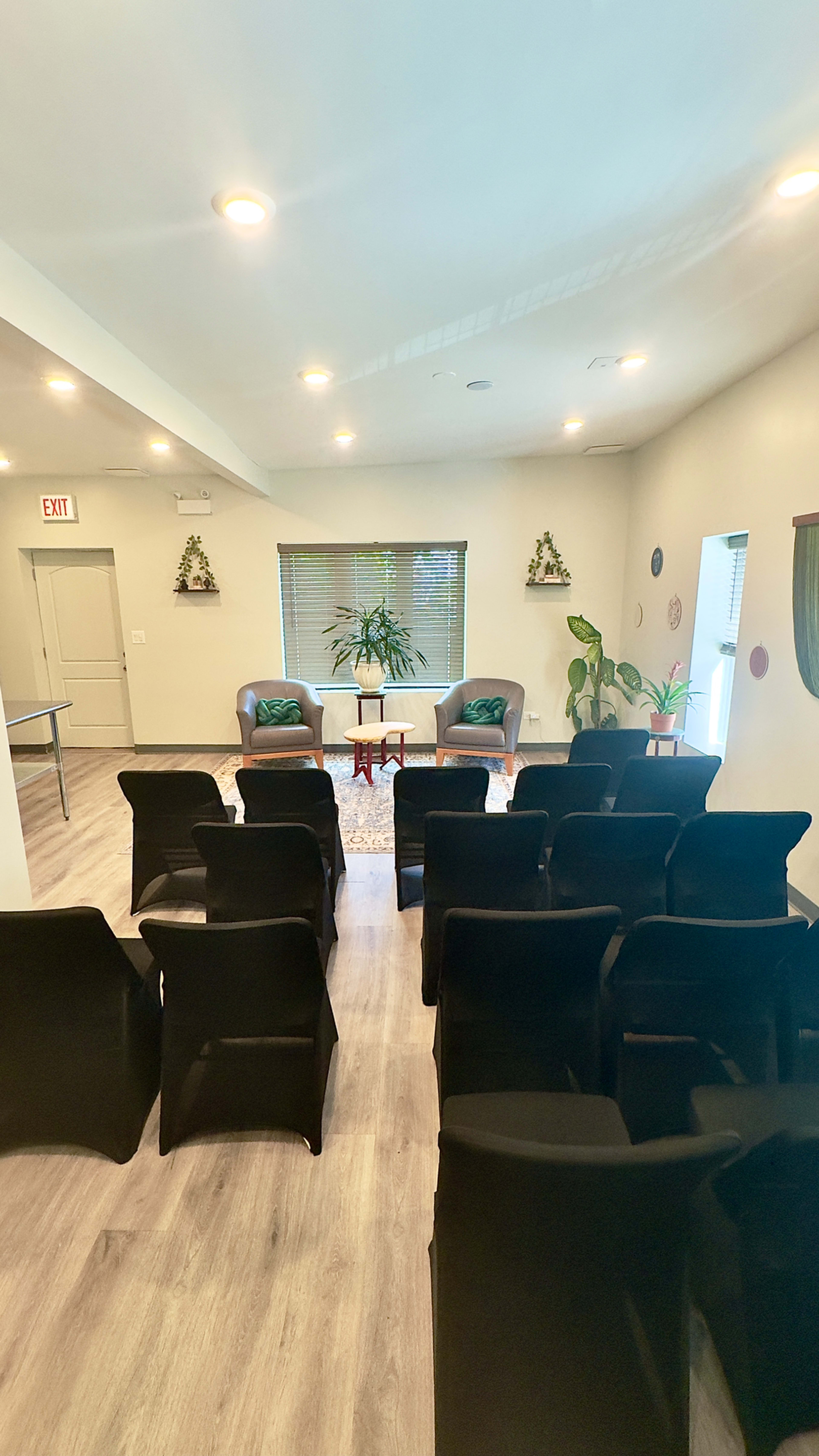 The image shows a light-filled room set up for an event, featuring two chairs and several rows of black chairs facing them, with plants and décor on the walls.