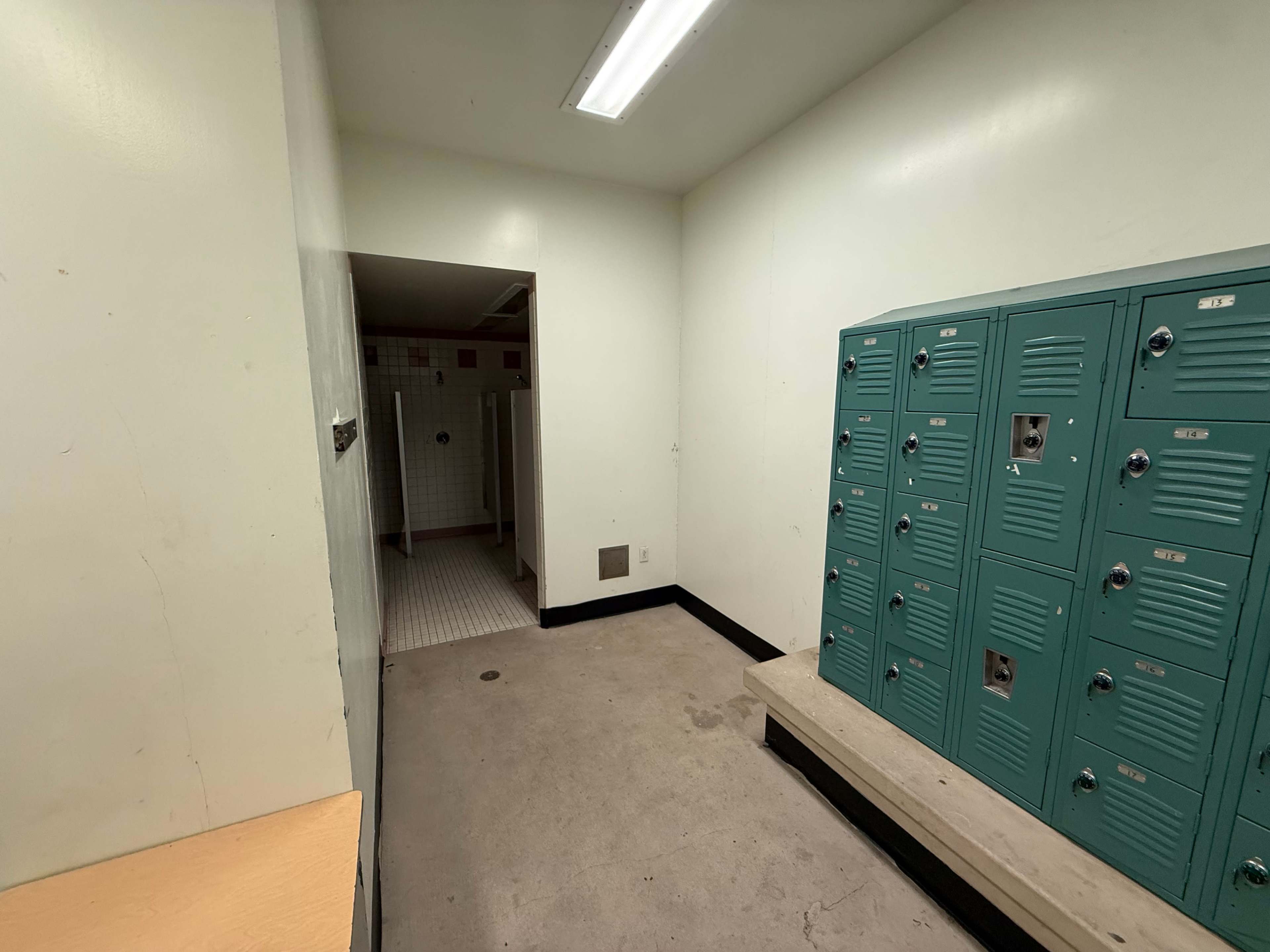 The image shows a small locker room with teal lockers on one side and a clean white tiled area that appears to have shower stalls at the back.