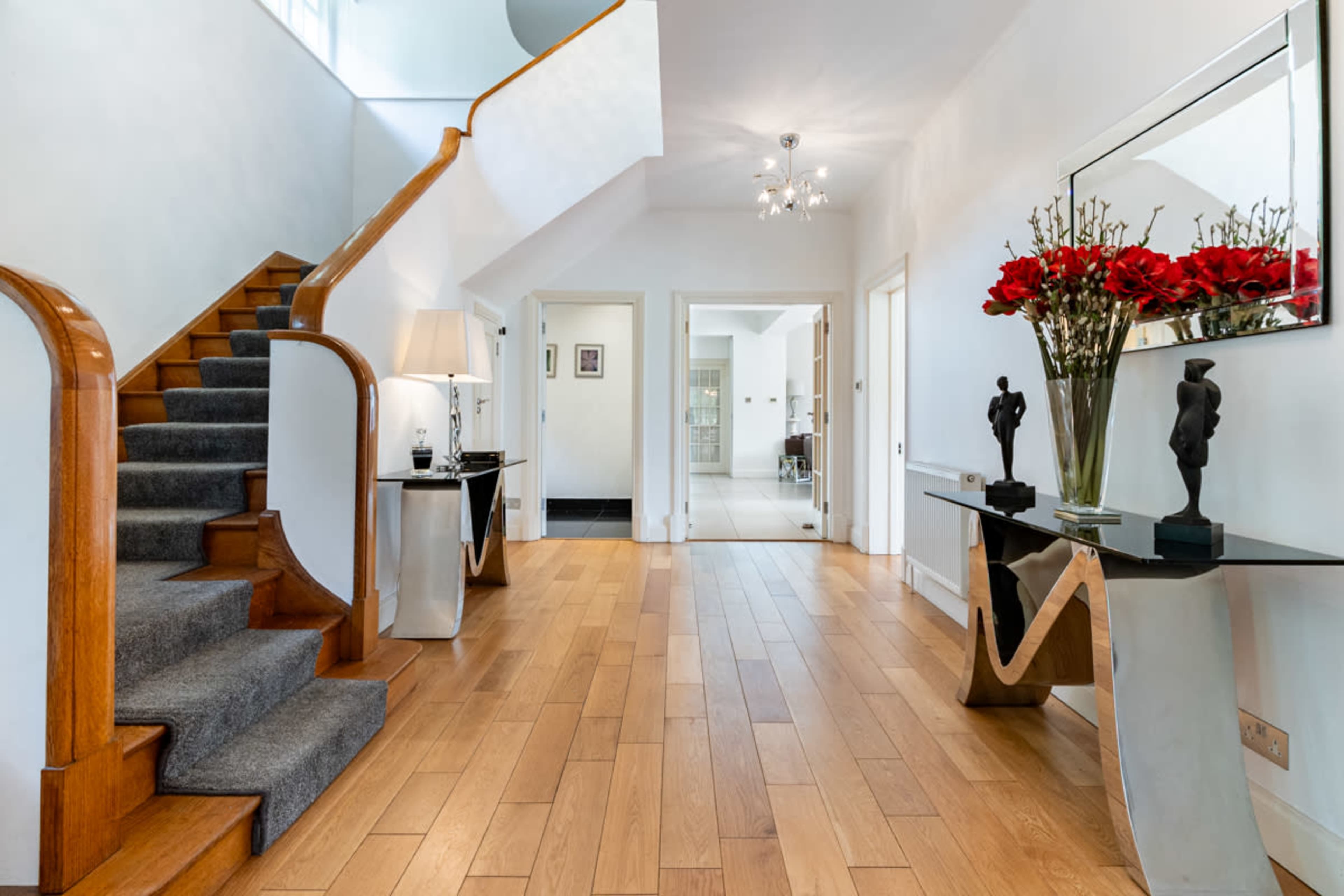 A well-lit entryway features a wooden staircase, a modern console table with decorative flowers, and a pair of sculptures on each side.