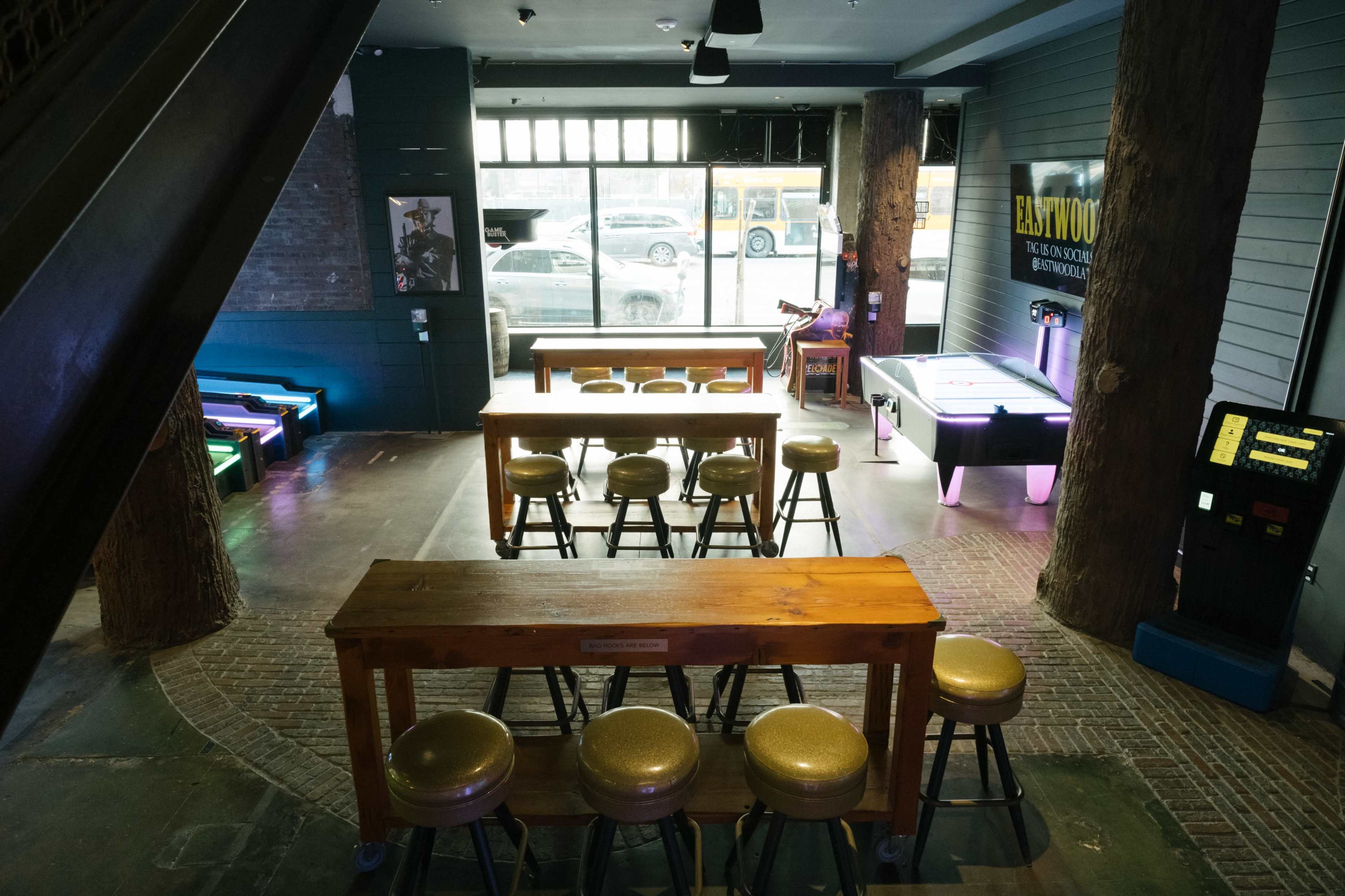 The image shows a spacious entertainment venue with wooden tables and stools, an air hockey table, and a vintage jukebox under dim lighting.