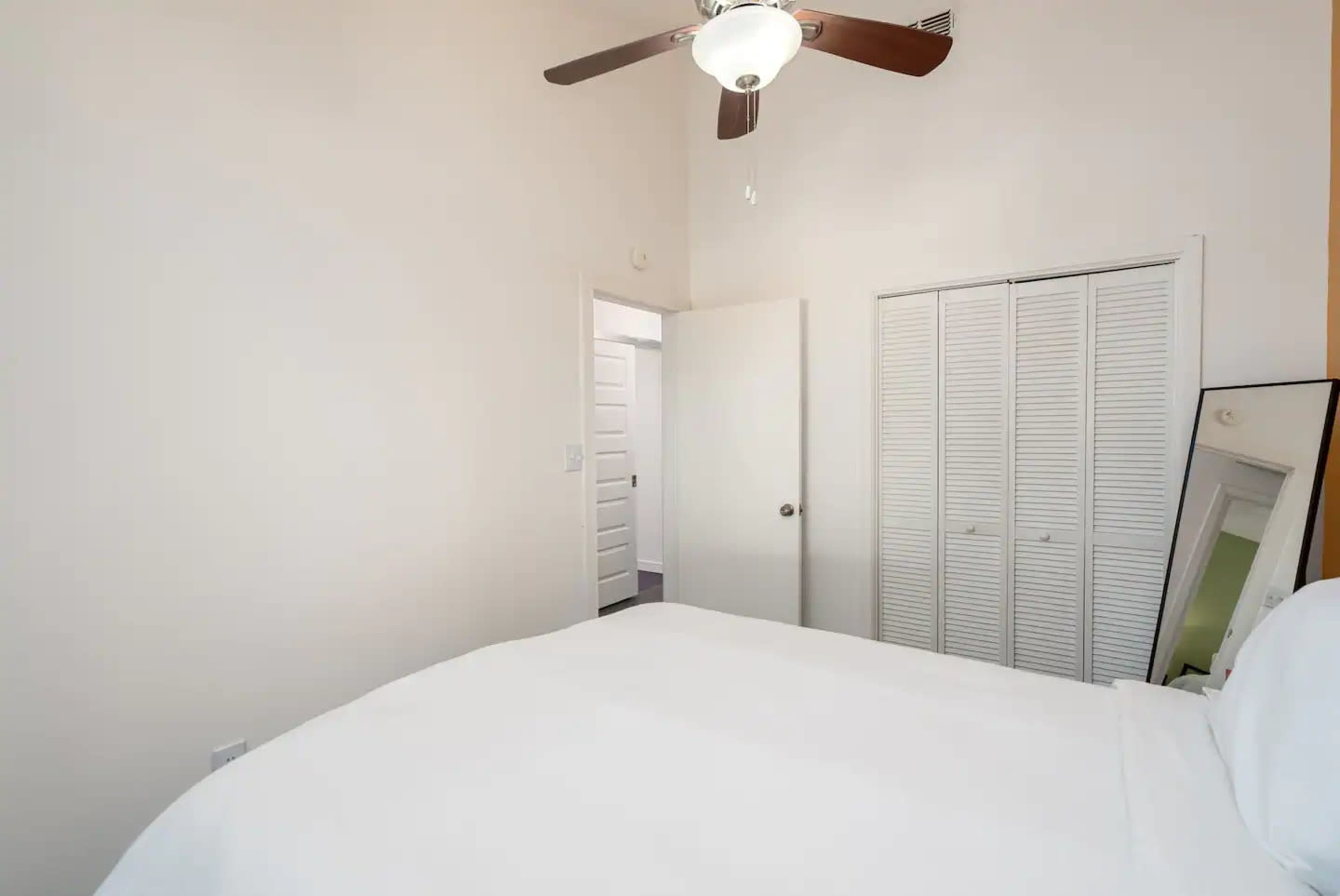 The image shows a bedroom with a bed, a ceiling fan, a closet with louvered doors, and a mirror reflecting part of the room.