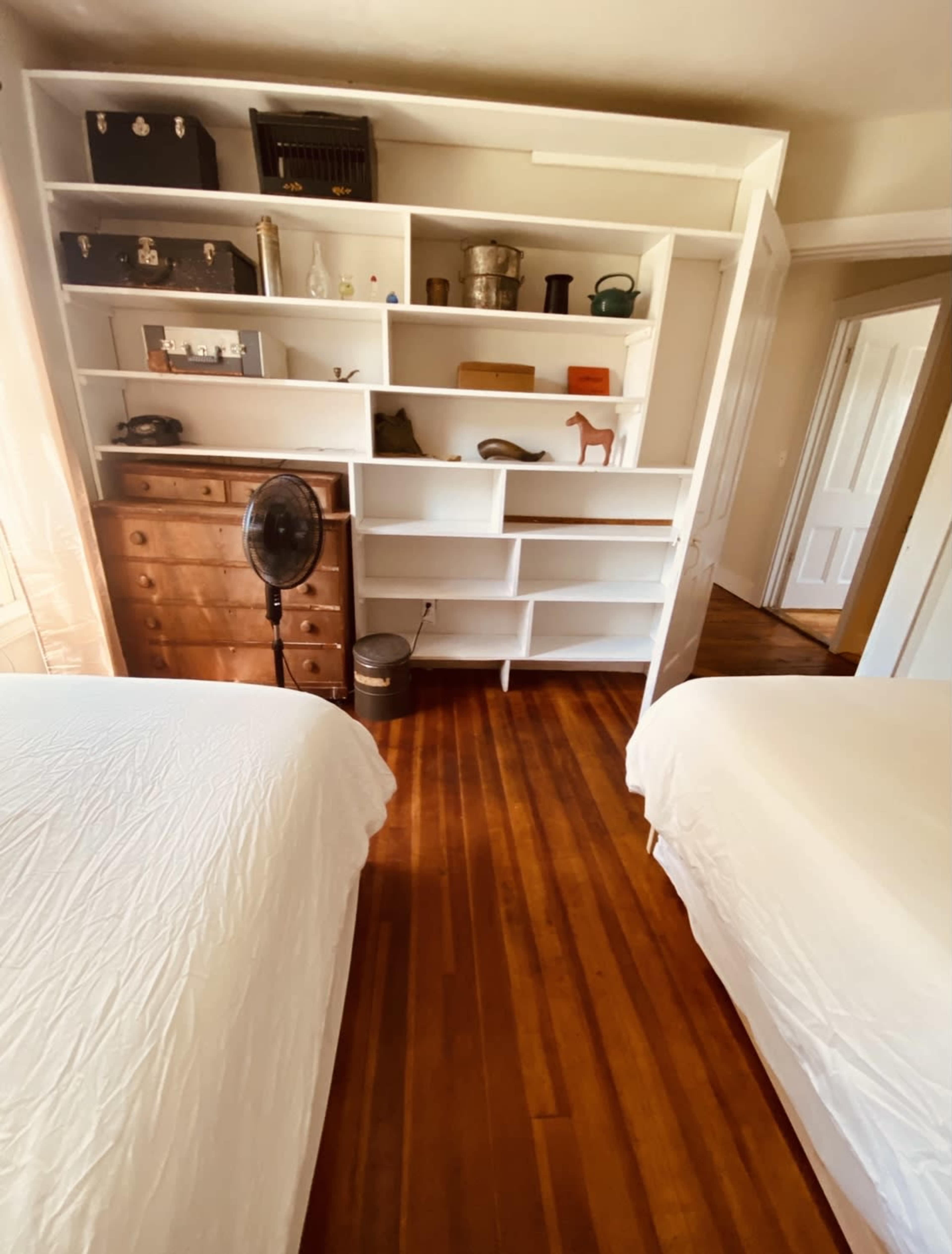 A bedroom with two beds, wooden flooring, and a white shelving unit displaying various objects.