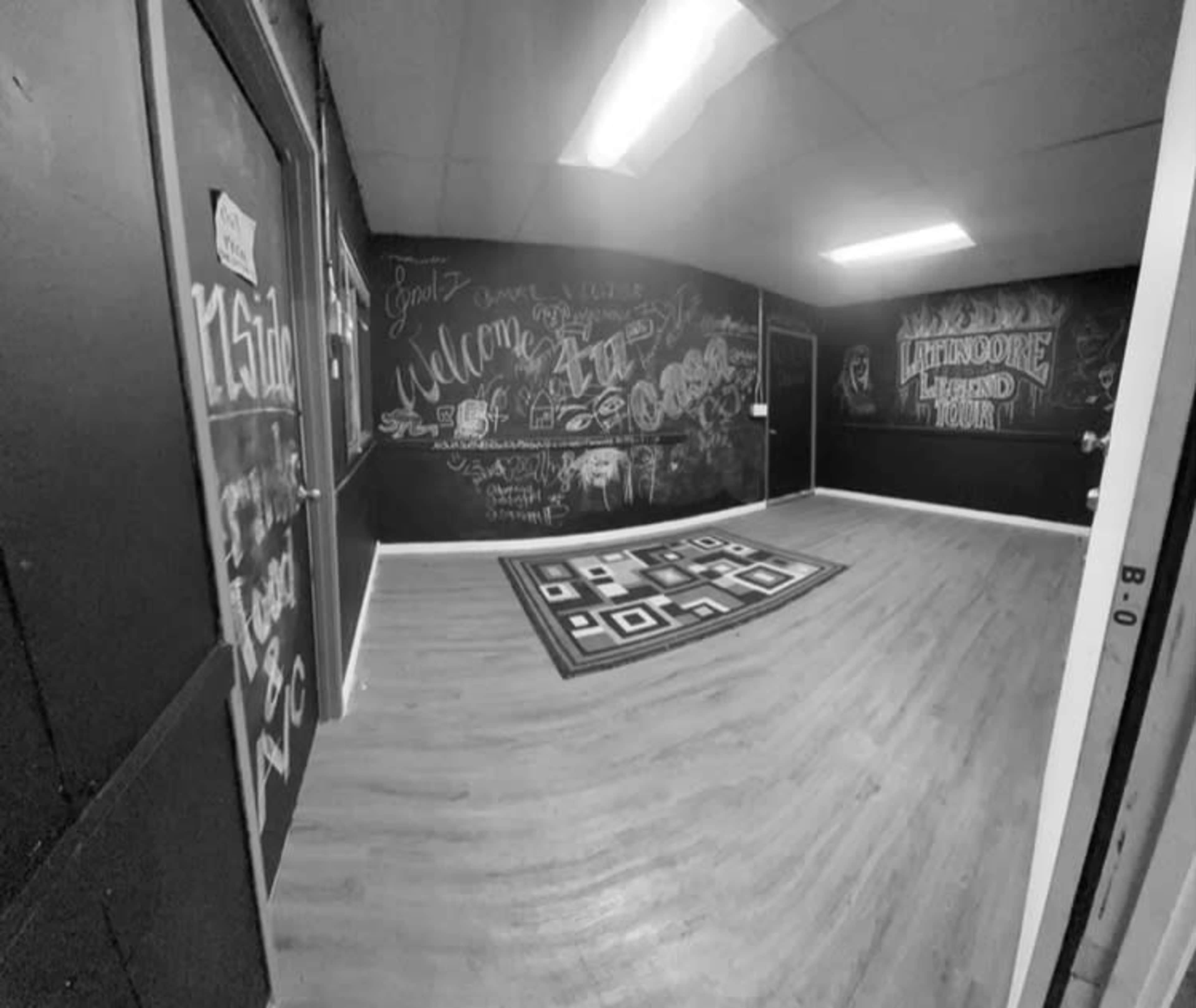 The image depicts a sparsely furnished room with black chalkboard walls covered in colorful graffiti and a geometric rug on the floor.