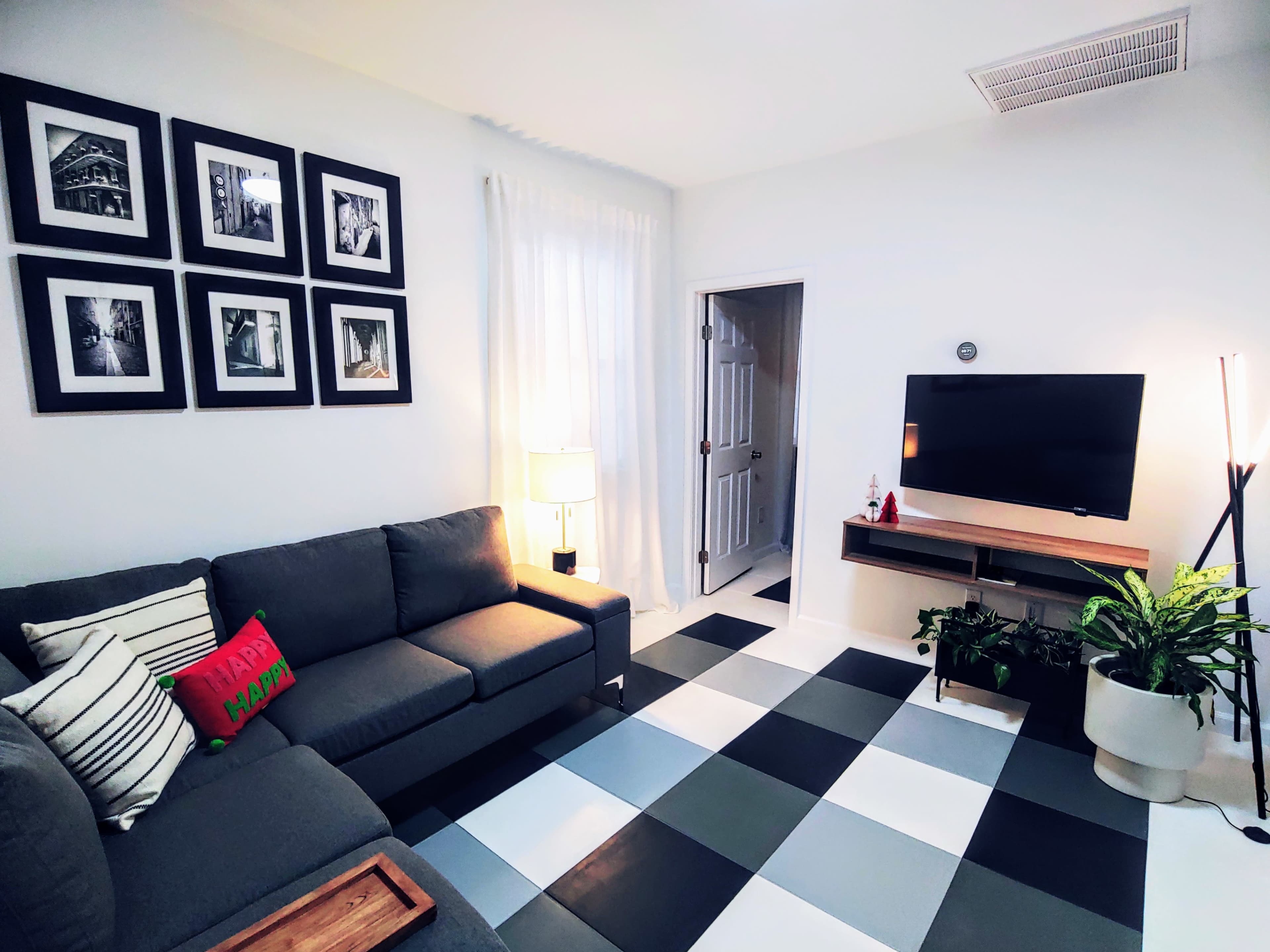 A modern living room features a gray sectional sofa, a wall with framed black and white photographs, a television mounted on a wooden stand, and a checkered floor in black, white, and gray.