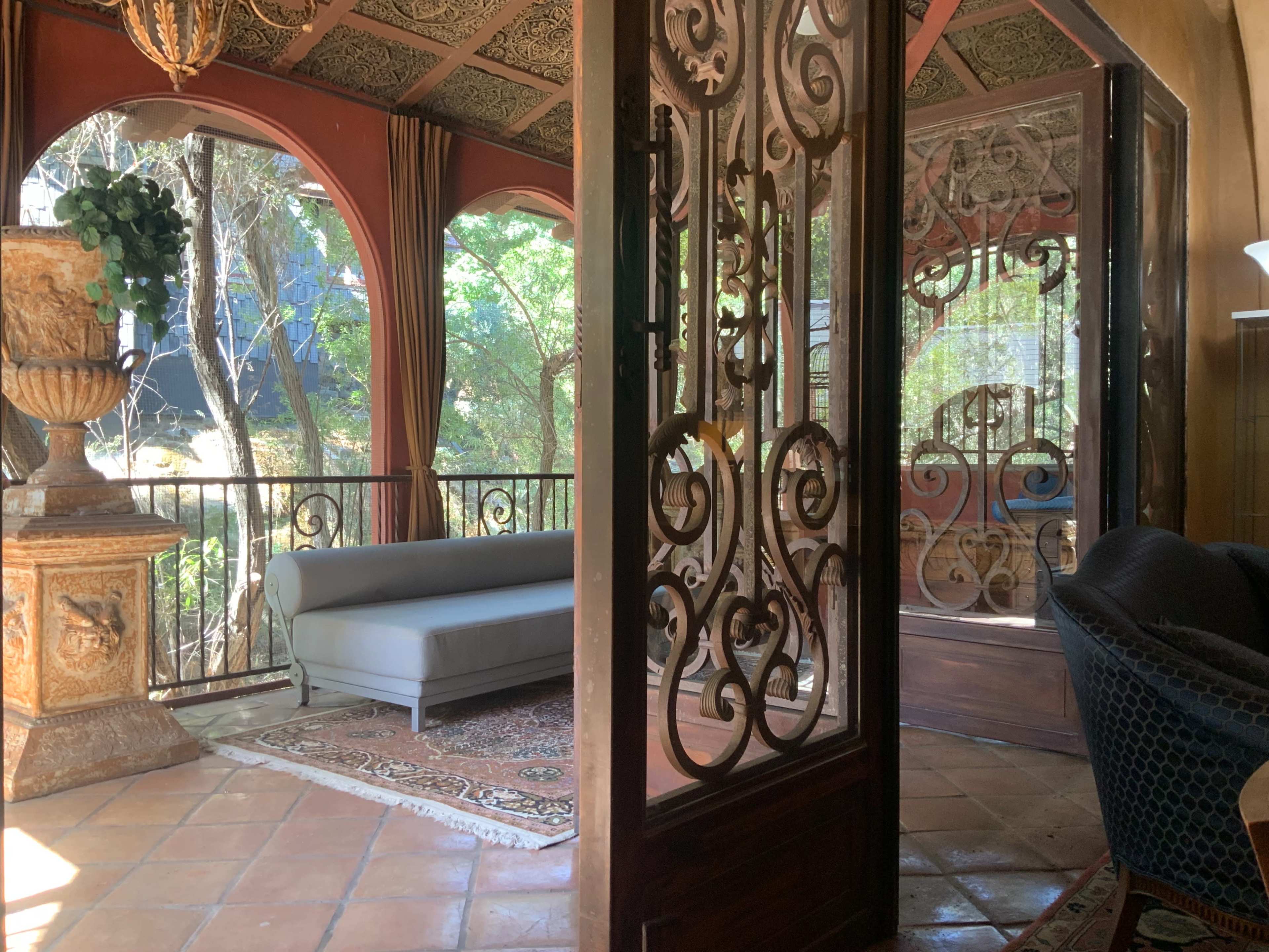 The image shows a room with ornate iron doors leading to a balcony, featuring a gray sofa, decorative vase, and a patterned rug on a tiled floor.