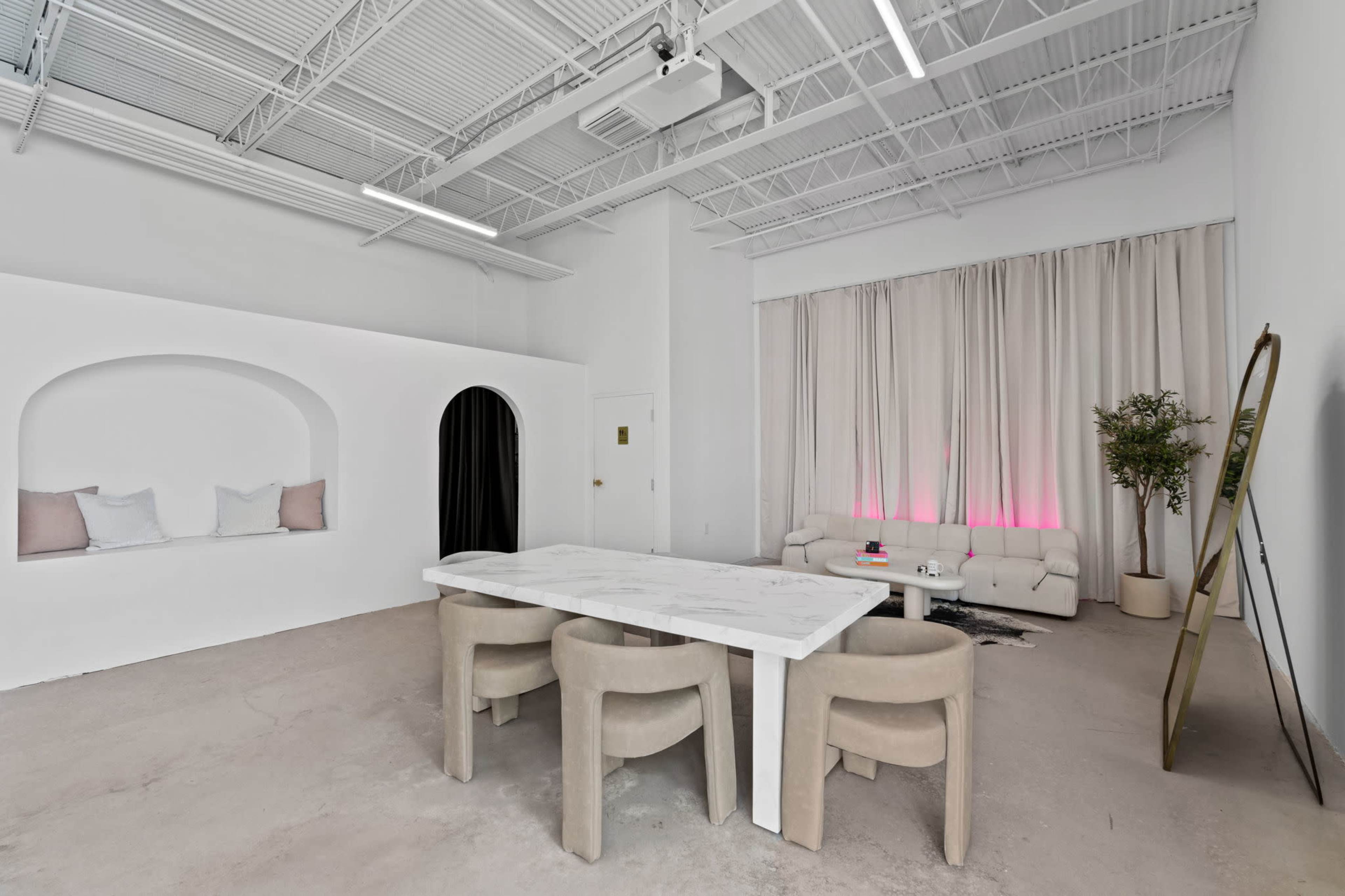 The image shows a spacious interior featuring a white marble dining table surrounded by four beige chairs, with a minimalist seating area and decorative elements along the walls.