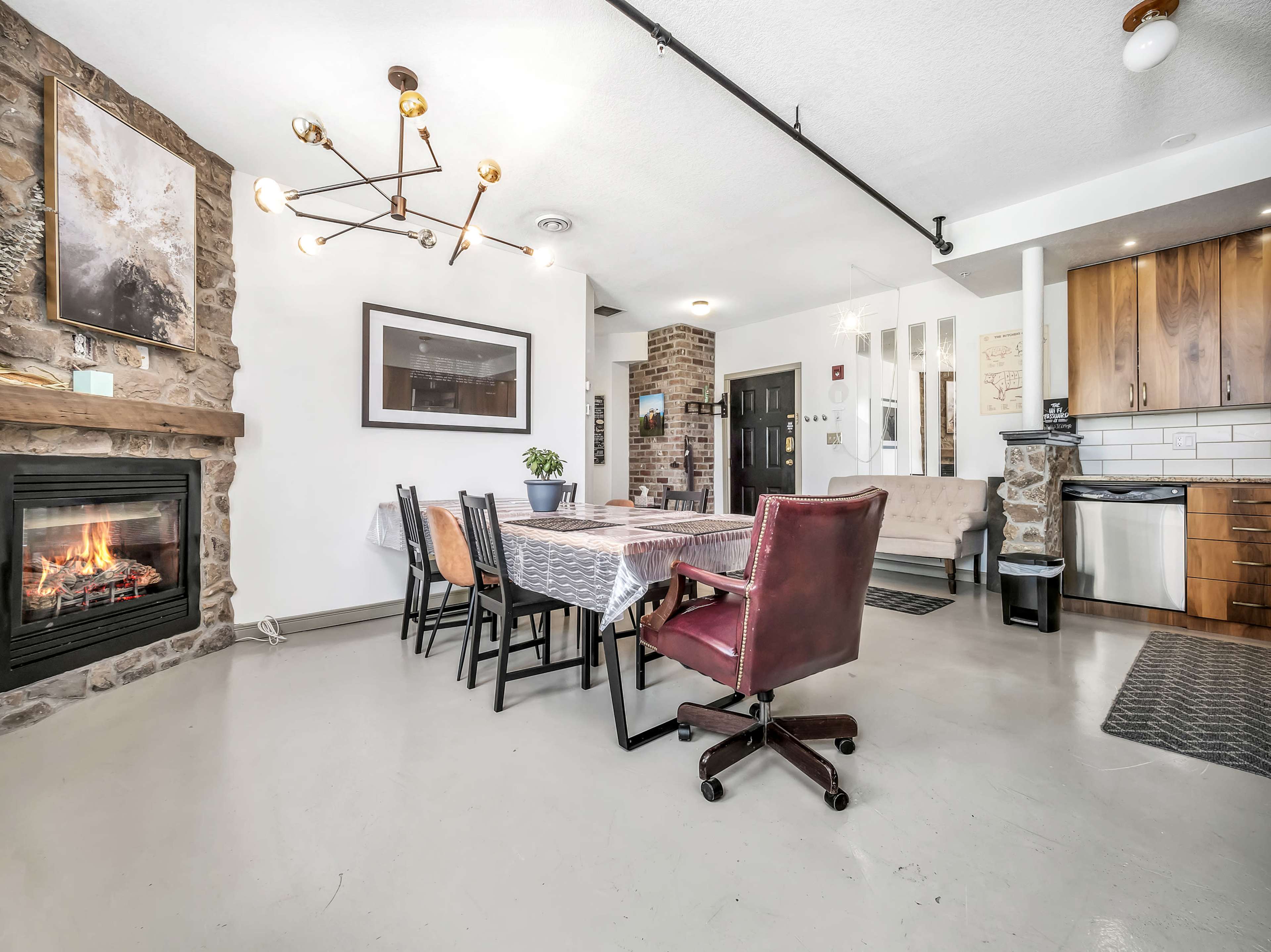 A modern, open-concept living space featuring a stone fireplace, a dining table set for several people, and a kitchen area with wooden cabinets.