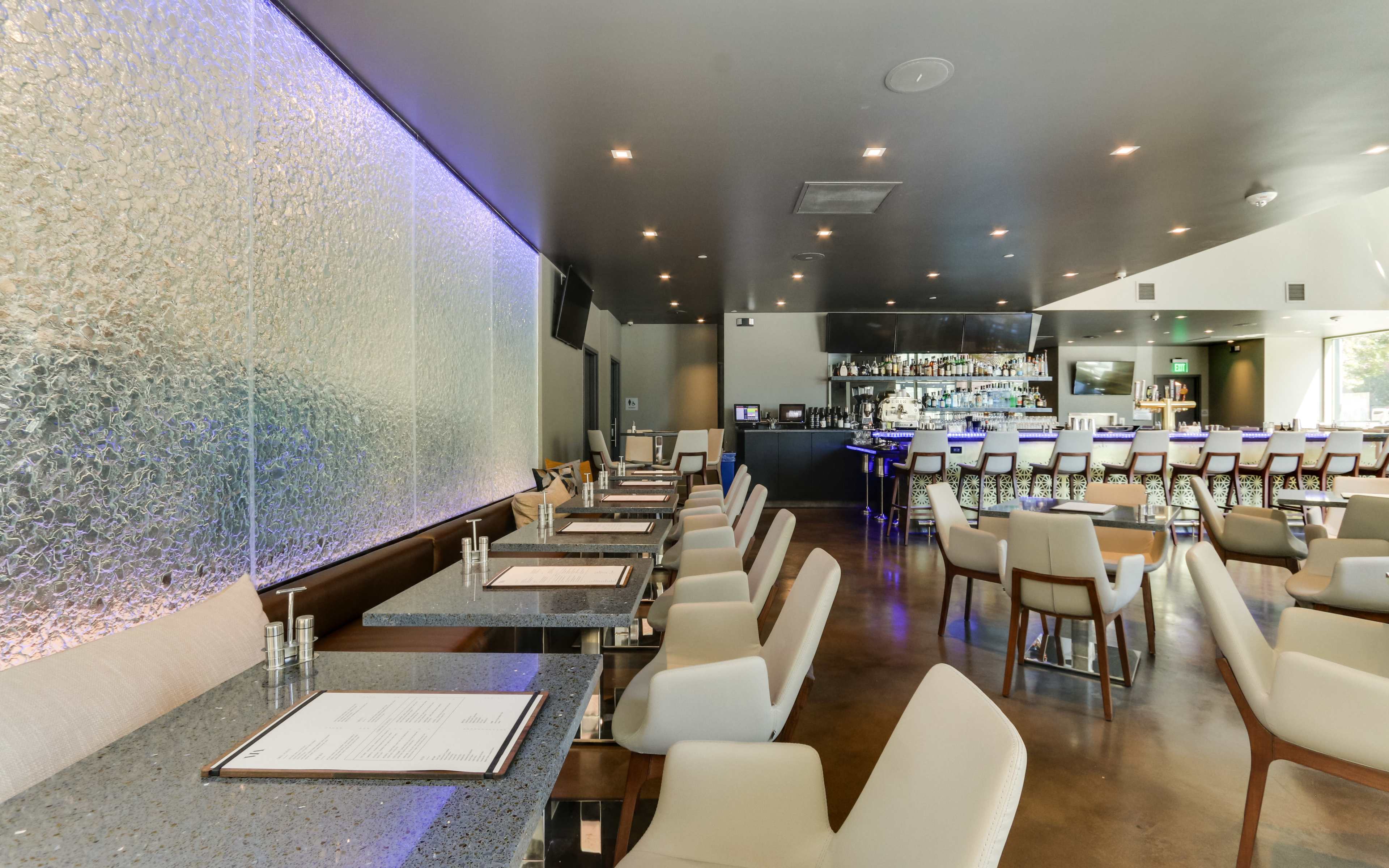 A modern restaurant interior with sleek tables, white chairs, and a bar area featuring illuminated shelves.