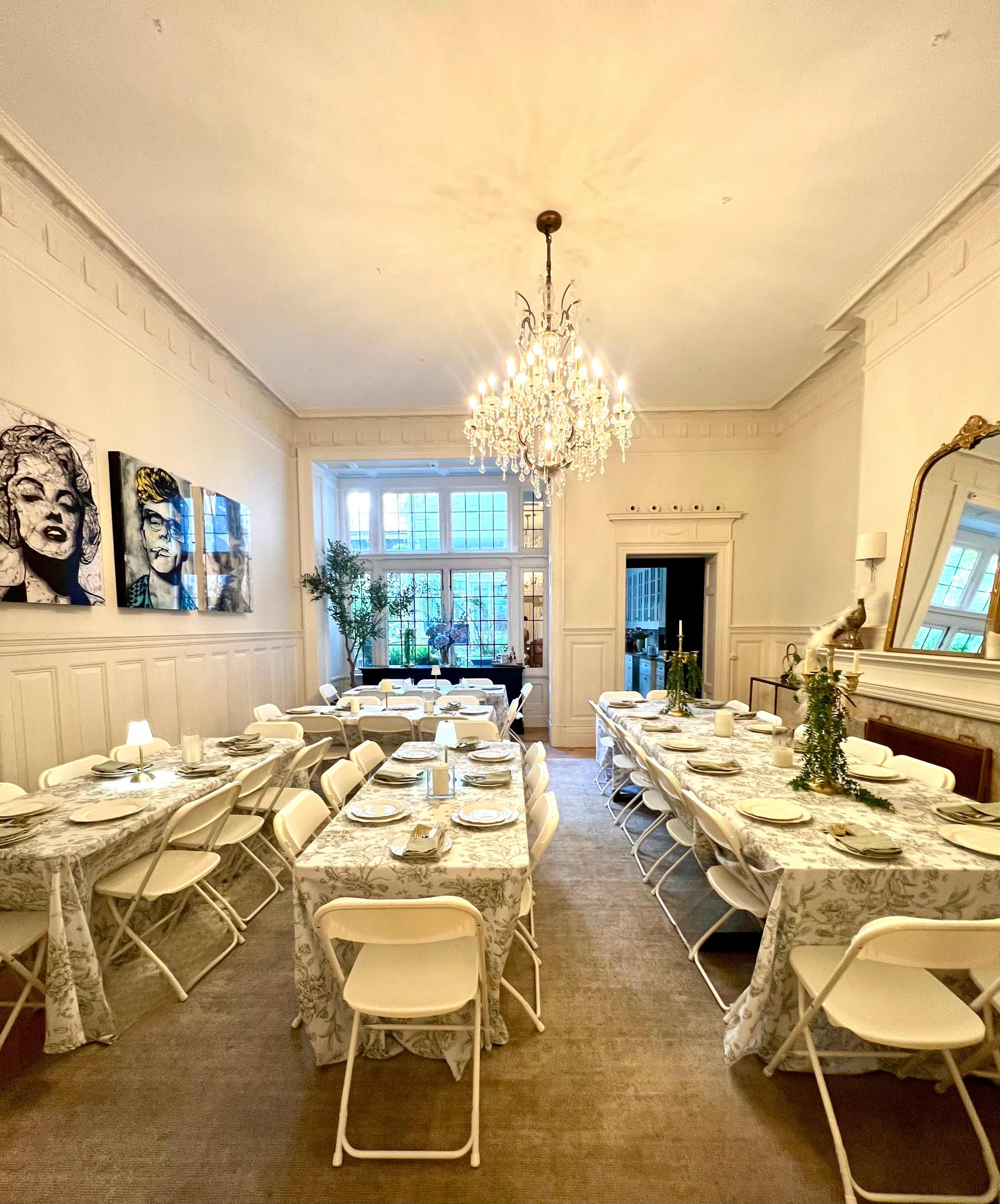 The image shows a spacious dining room with elegantly set tables covered with floral tablecloths, illuminated by a chandelier and featuring large windows.