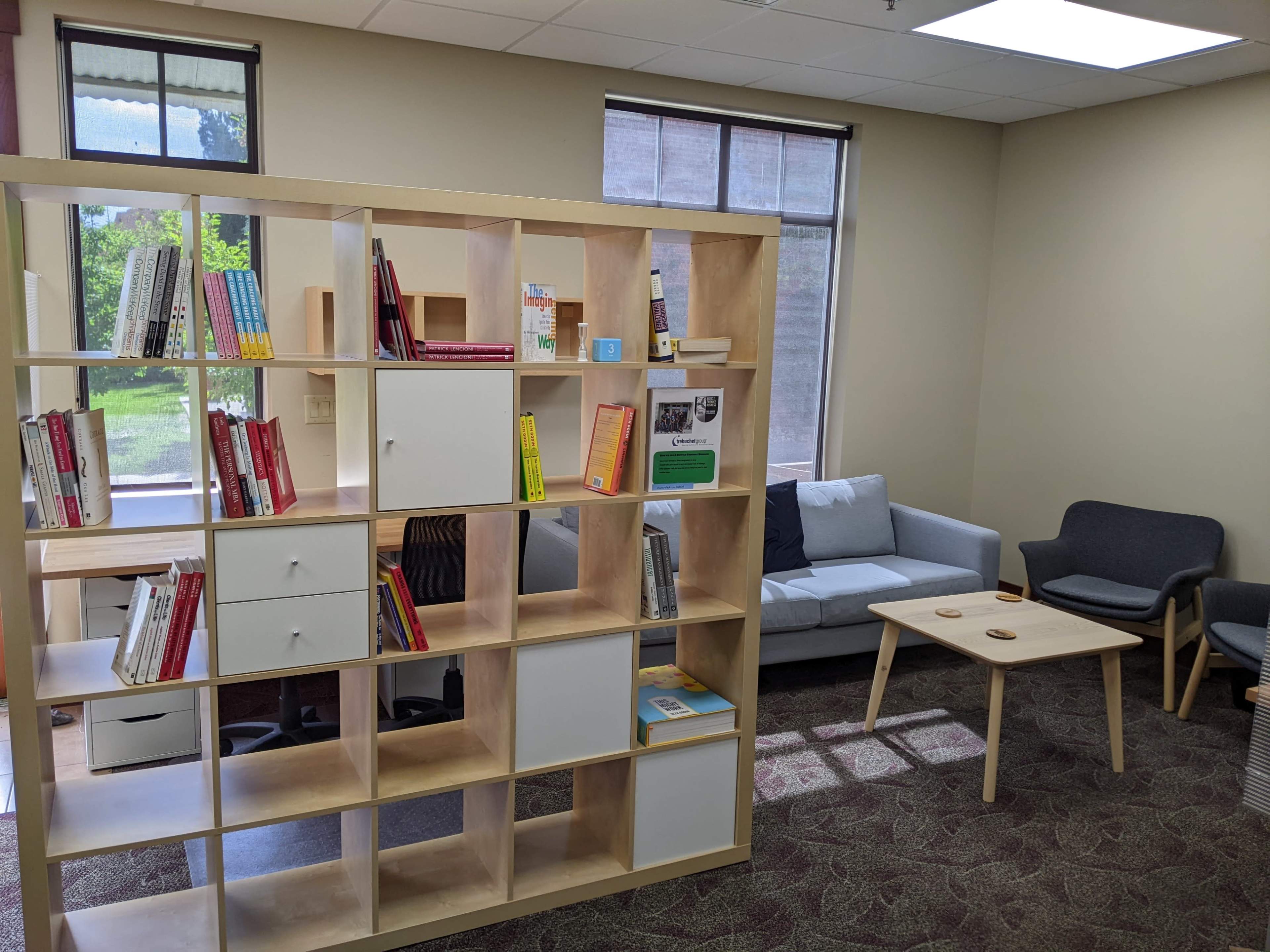 The image shows a modern office space with a bookshelf, a seating area featuring a gray couch and two chairs, and a table in front.